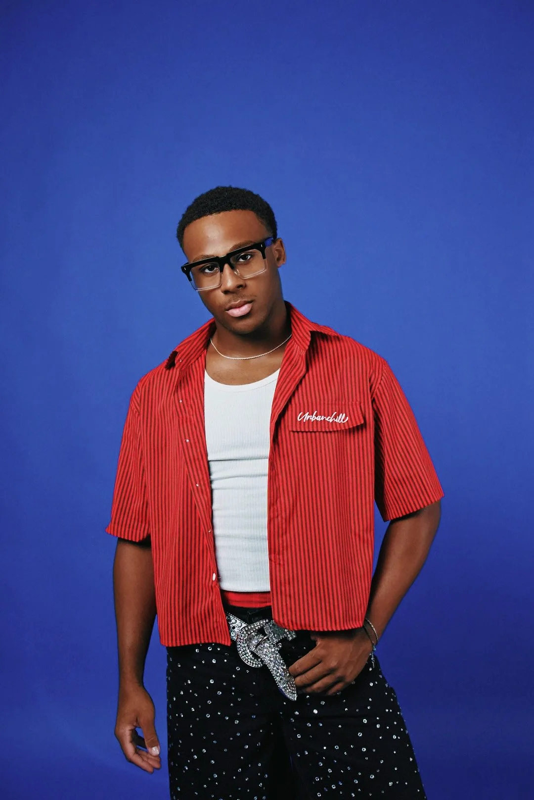 Young man in a red and black striped shirt over a white T-shirt, wearing glasses, black pants with white dots, and a large belt buckle, posing against a blue background.