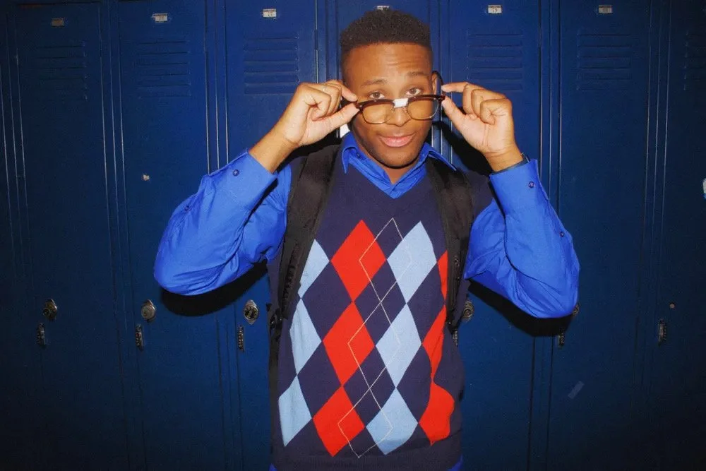 A young male student with glasses, wearing a blue shirt, a colorful argyle sweater vest, and a backpack, standing in front of blue lockers in a school hallway.