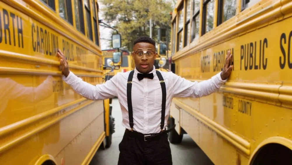 Young man in glasses, white shirt, black suspenders, and bow tie standing between two yellow school buses, holding onto each bus.