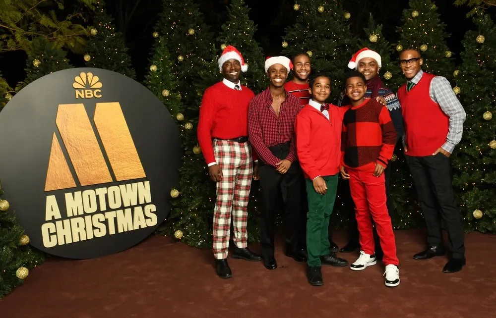 Group of seven people, six men and one woman, dressed in festive Christmas attire, standing in front of decorated Christmas trees, with a large sign reading 'A Motown Christmas' and the NBC logo.