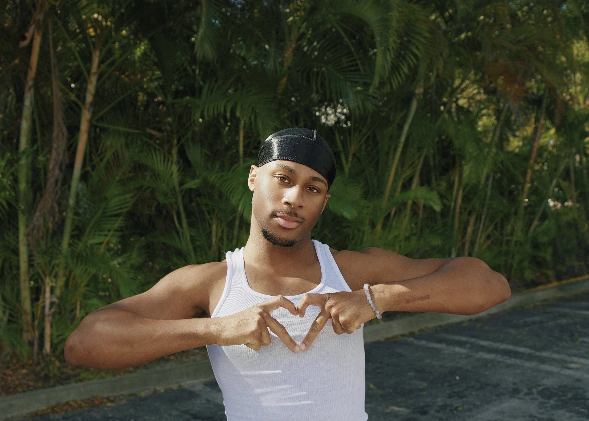 Young man with a black head wrap and a white tank top making a heart shape with his hands in front of green foliage.