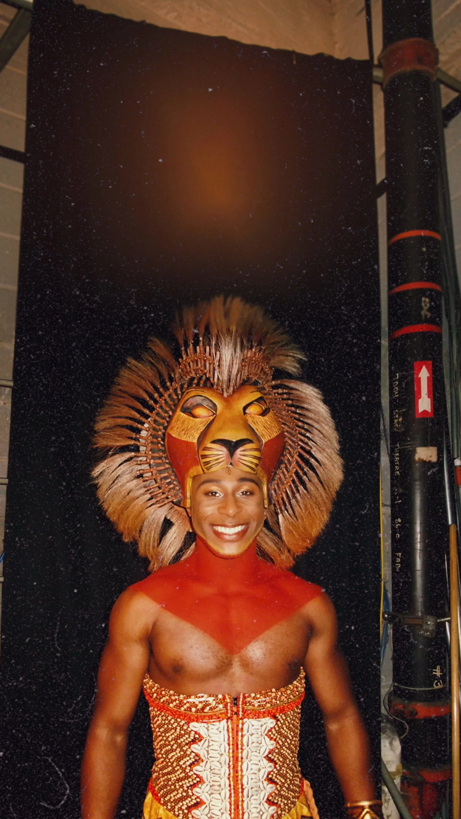 A woman with dark skin and a bright, joyful smile is dressed in traditional African dance attire. She wears an elaborate lion mask with a golden face and a large, multicolored mane made of feathers. Her outfit includes a red and white patterned skirt and her body is painted red around her neck and shoulders.