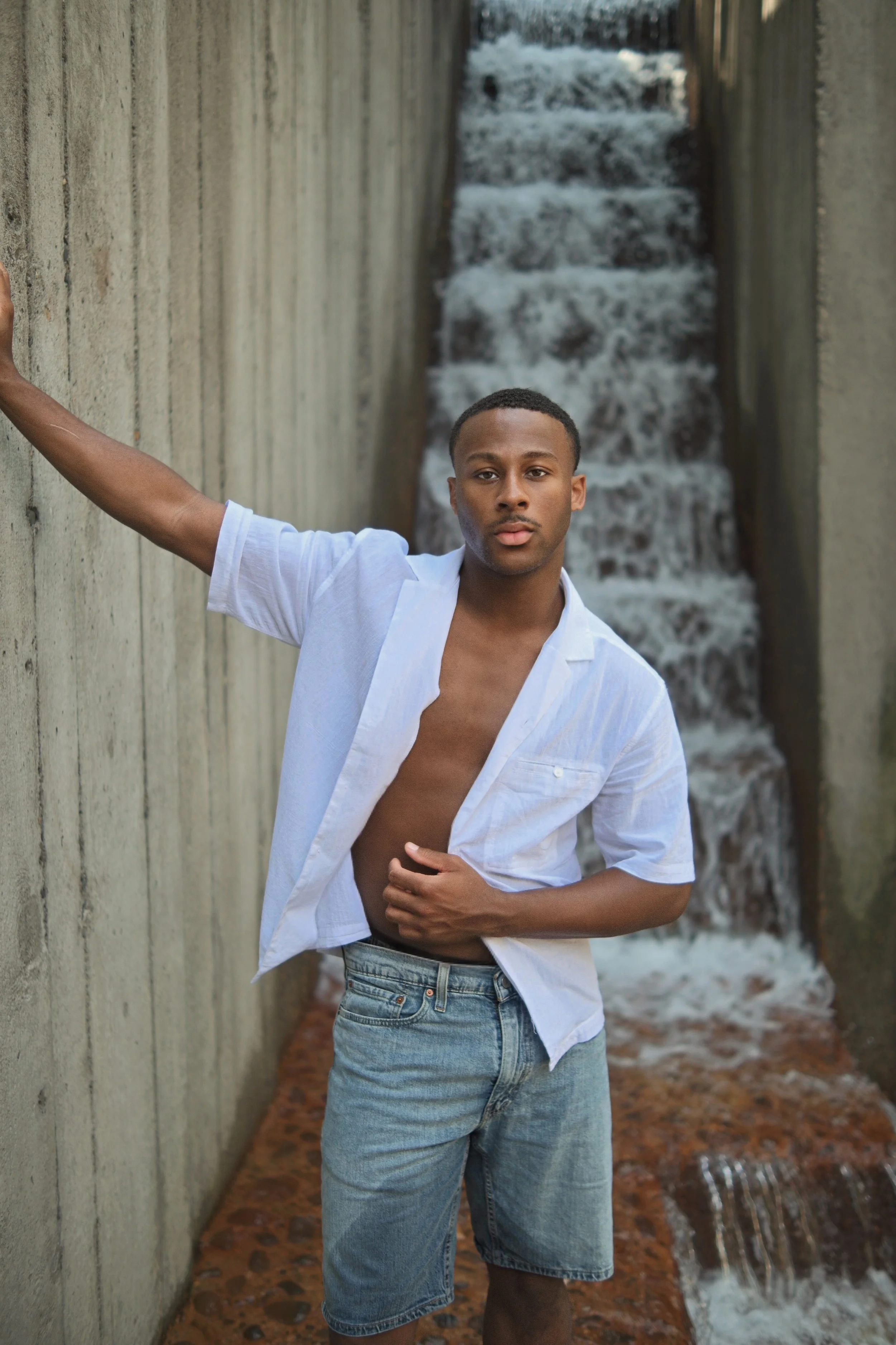 A young man with short hair and medium skin tone, wearing a white shirt partially unbuttoned and blue jeans, standing against a concrete wall with a waterfall in the background.