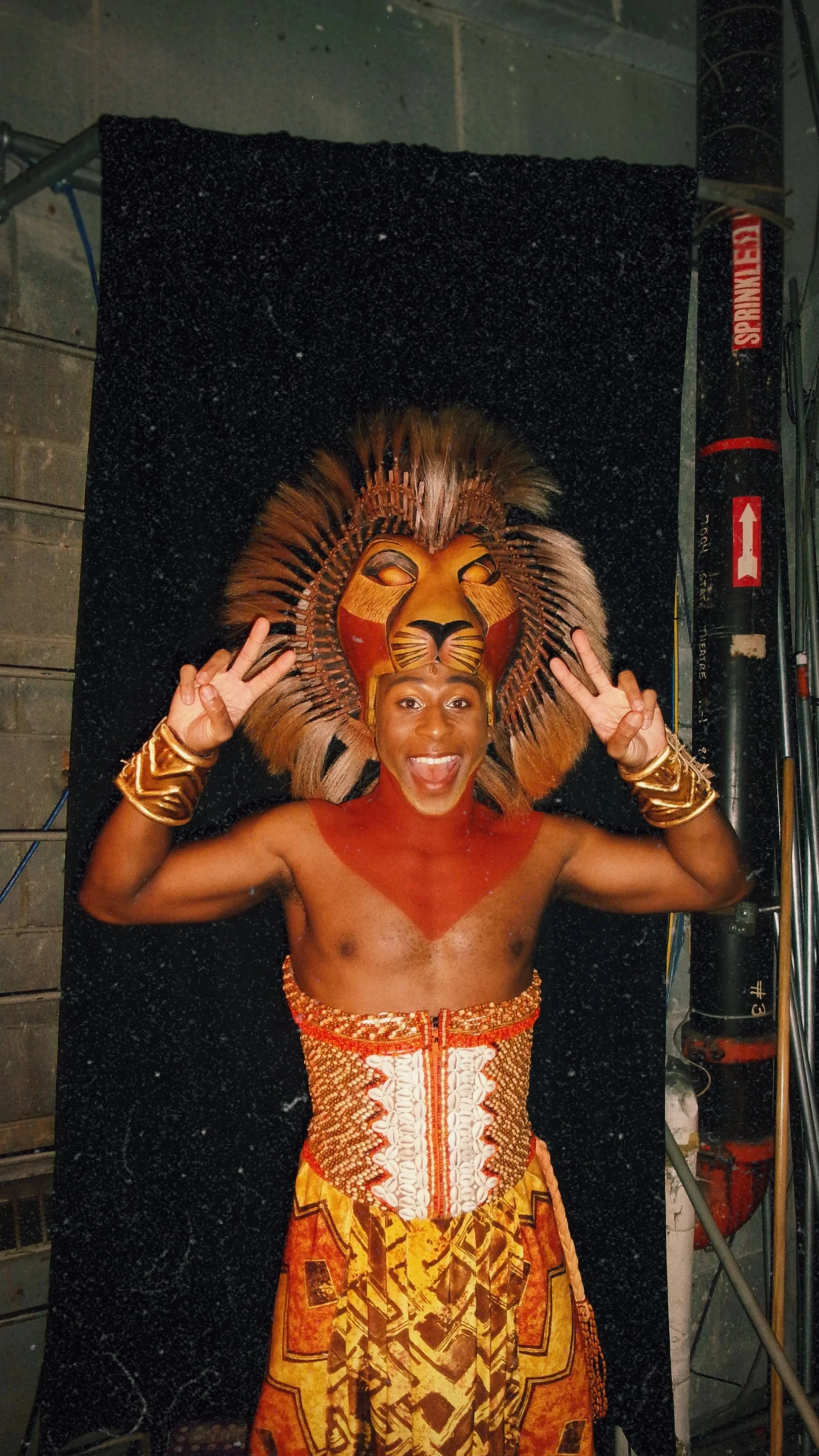 A performer with a lion mask and traditional African-inspired costume making a peace sign with both hands.