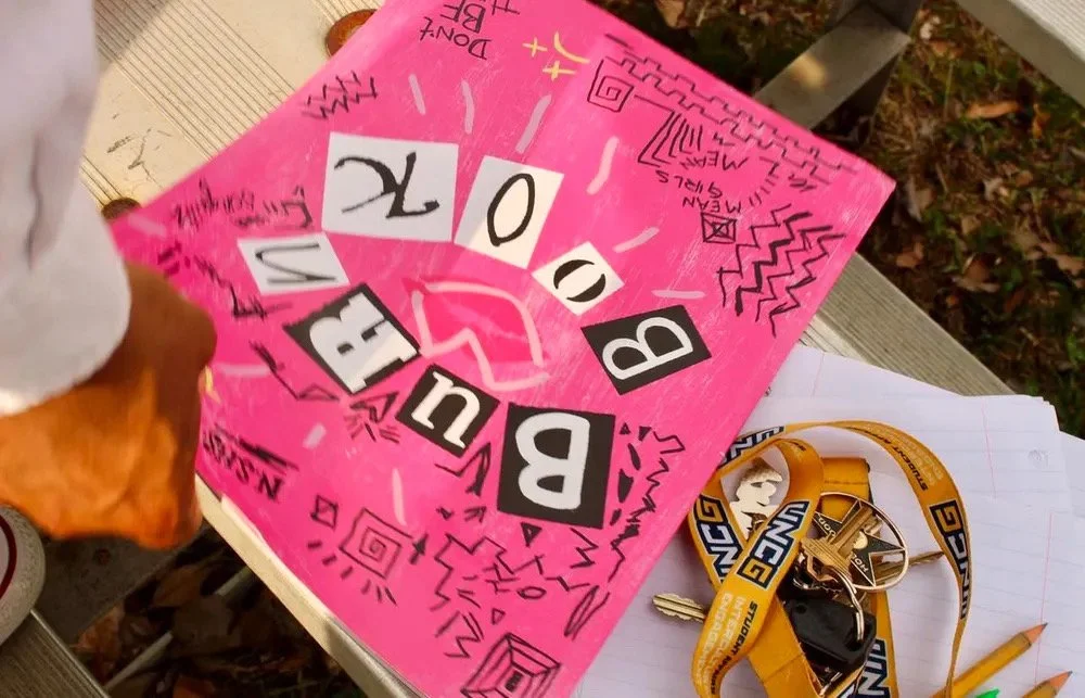 A pink decorated sign that says 'BIRTHDAY' and '10', with doodles and drawings. A white and yellow LUMS lanyard with keys and a black ID badge, and some orange pencils placed on lined paper.