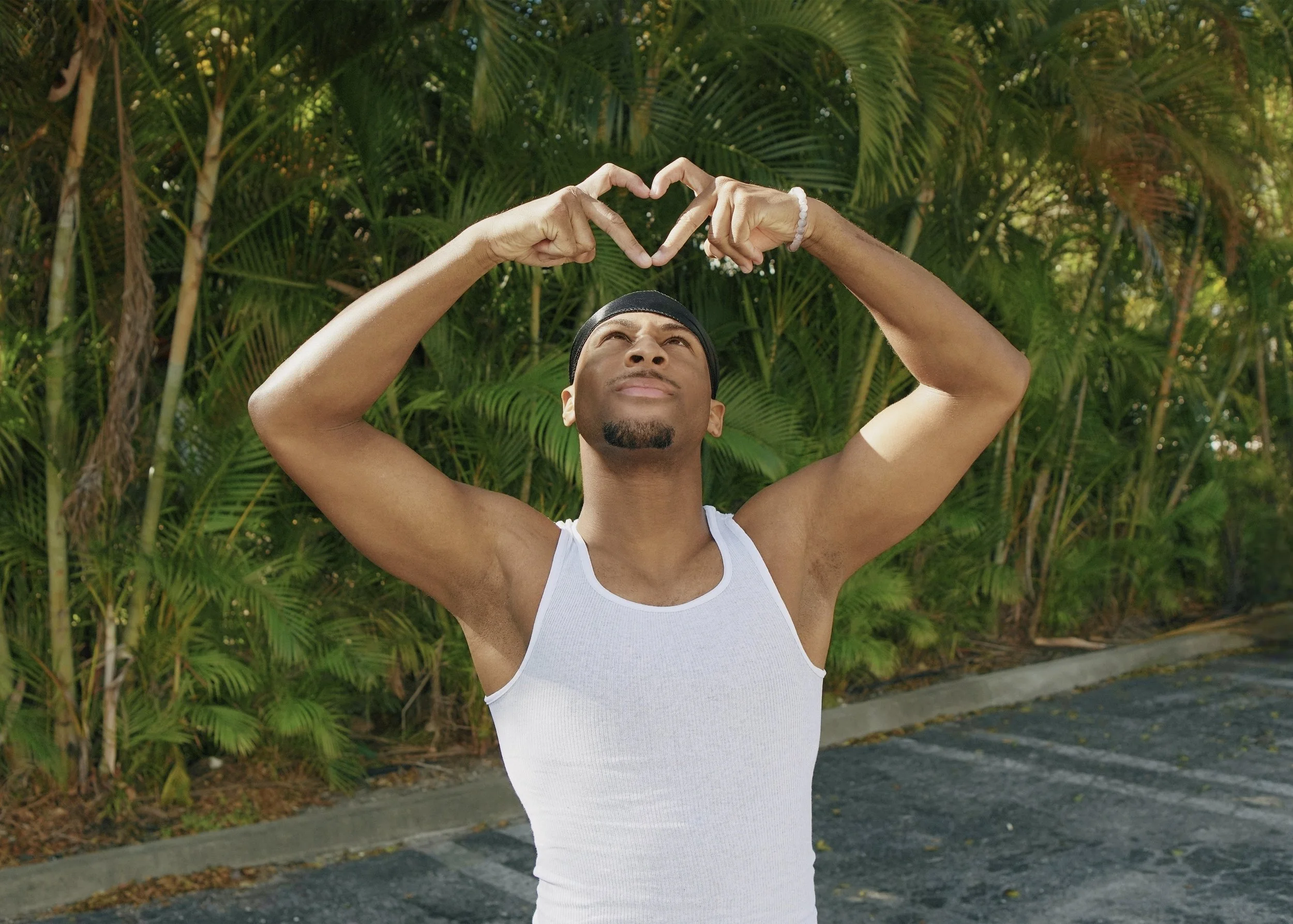 A man wearing a white tank top and a black headband standing outdoors with green tropical plants in the background, forming a heart shape with his hands above his head.