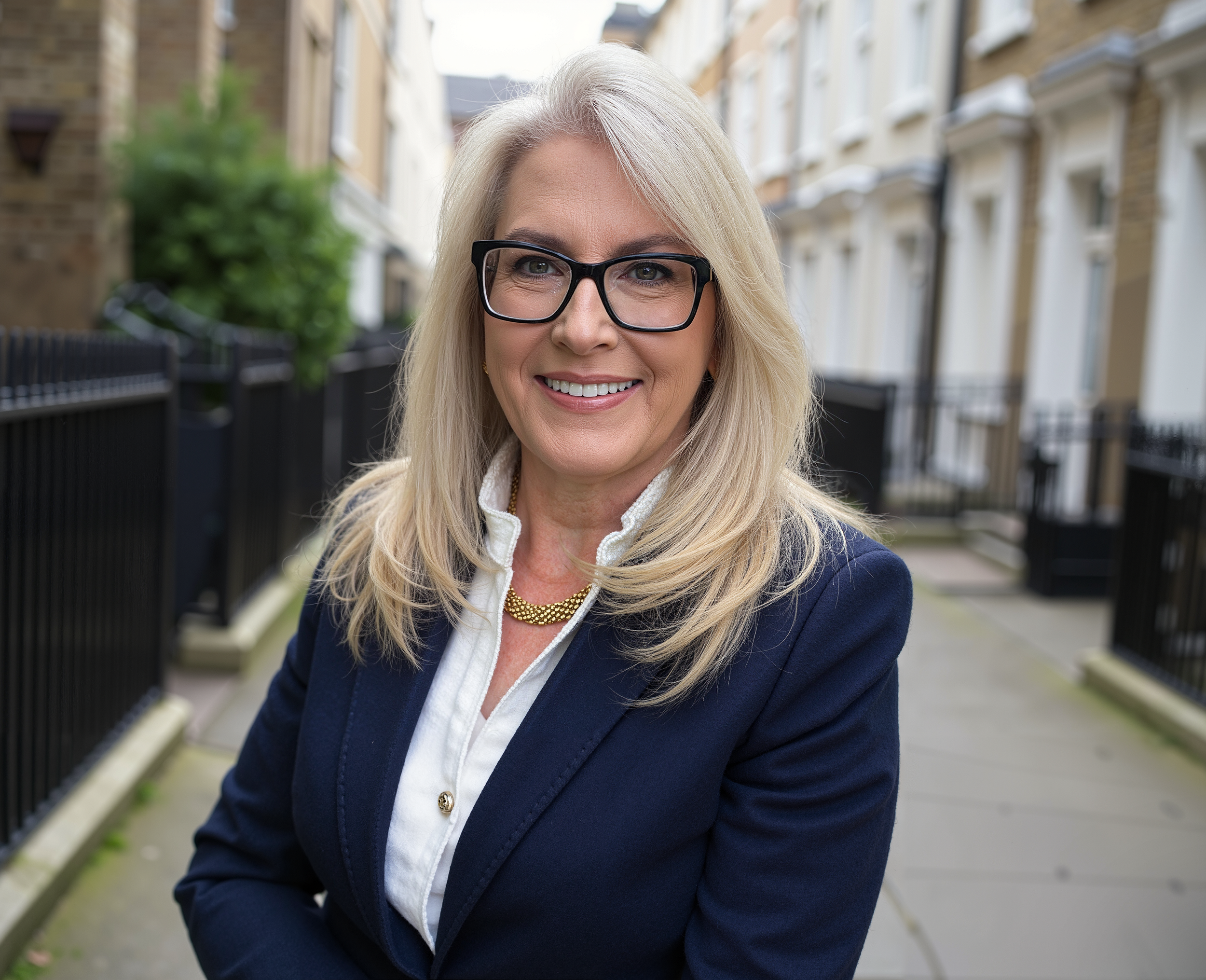 A blonde woman wearing glasses smiling in an outdoor urban setting with residential buildings and a walkway.