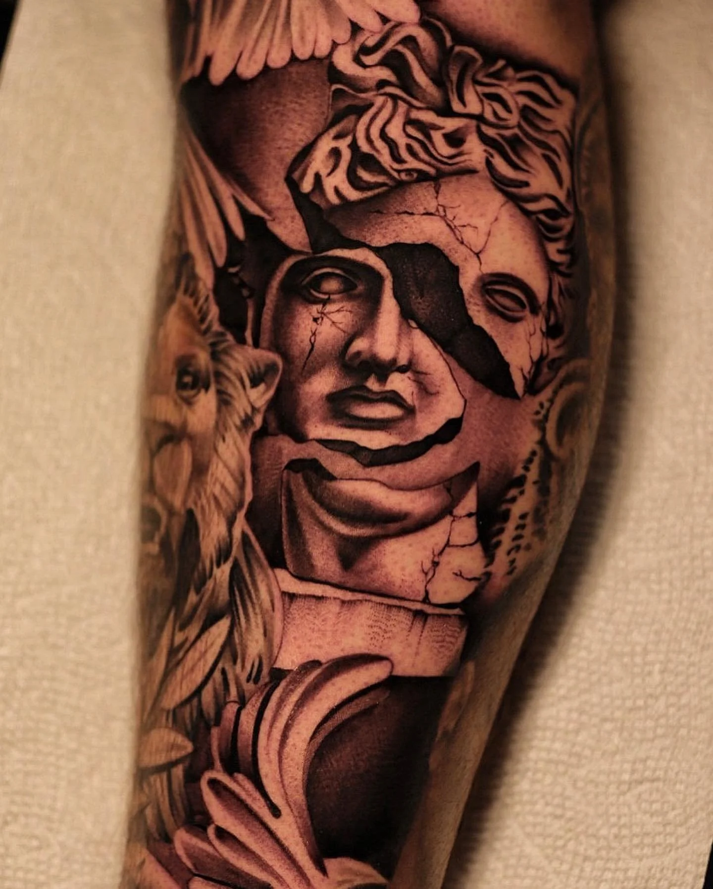 A detailed black and gray tattoo on an arm depicting a woman's face with cracks, a blindfold over one eye, surrounded by angel wings, a tiger, and floral elements.
