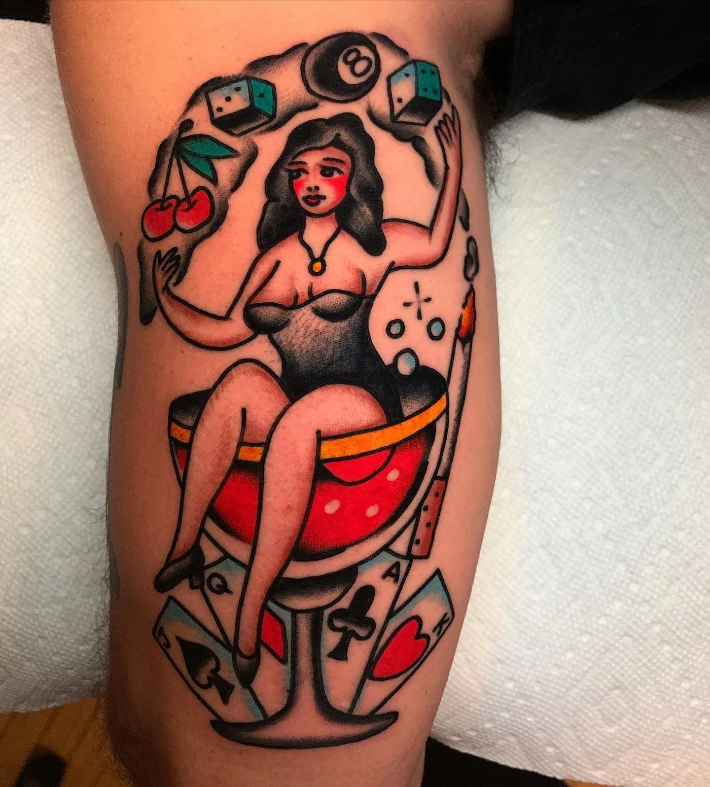 Tattoo of a woman with dark hair sitting on a roulette wheel, surrounded by dice, cherries, and playing cards, in a traditional tattoo style.