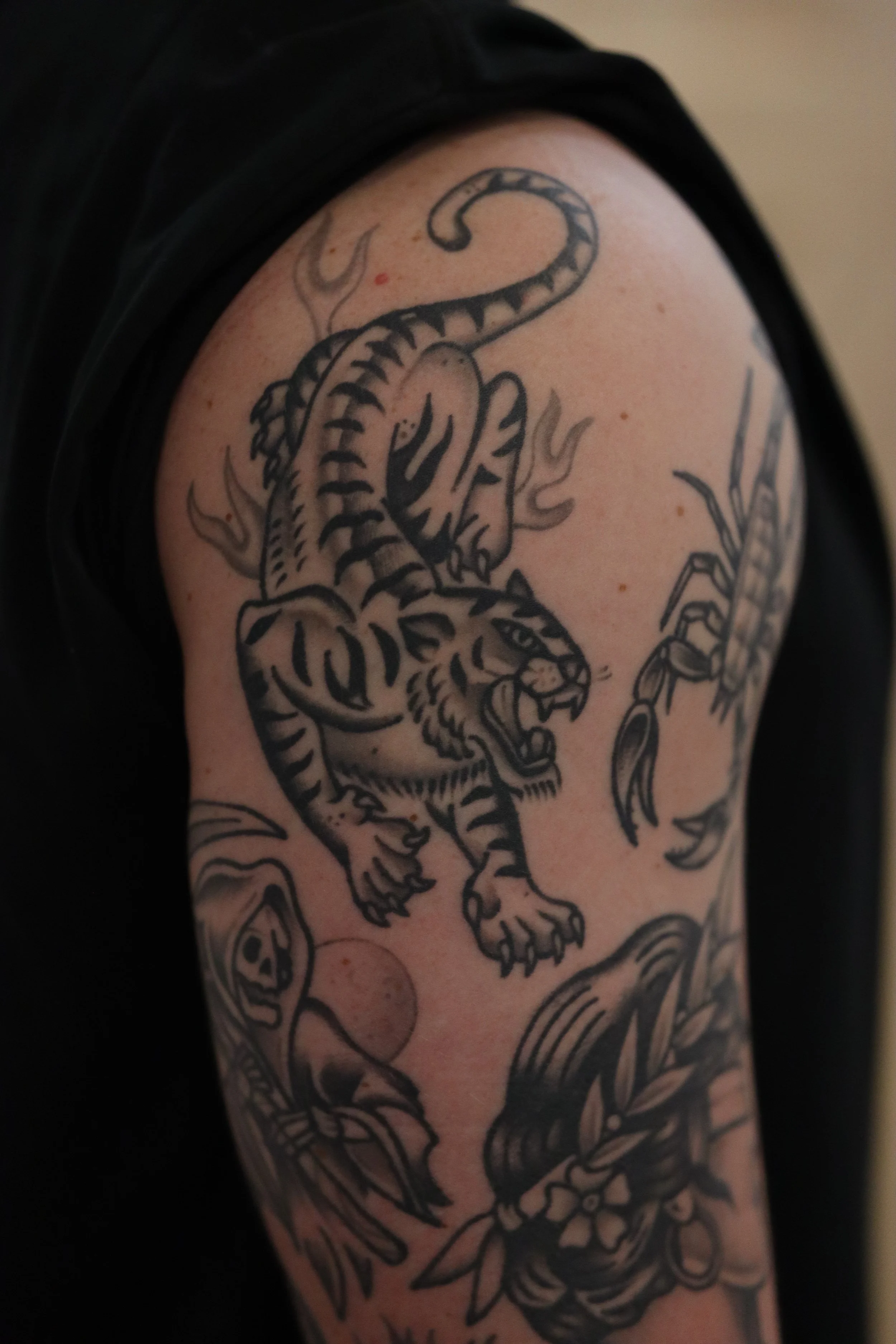 Black tattoo of a fierce tiger on a person's upper arm surrounded by flames and skulls.