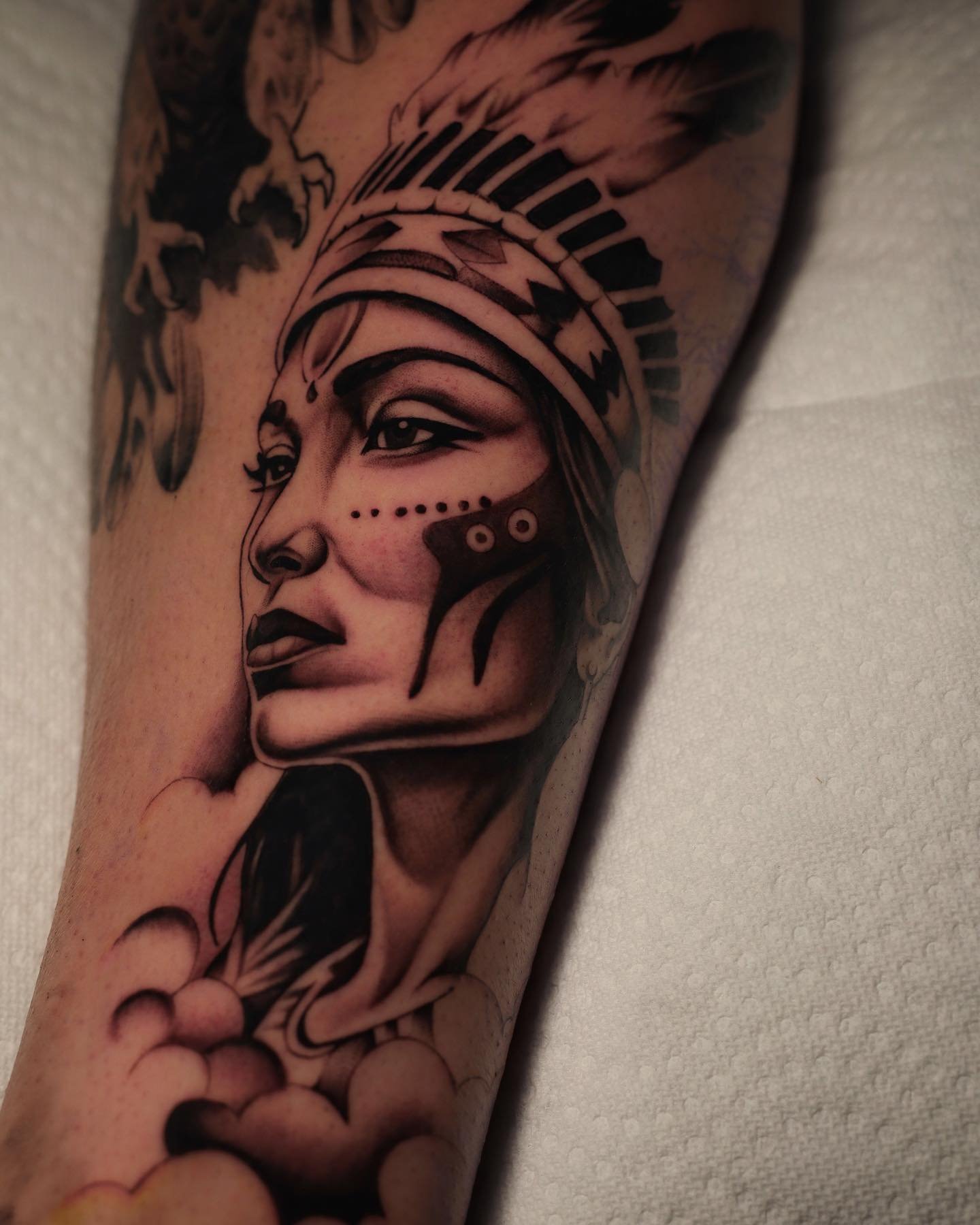 Black and gray tattoo of a woman's face with makeup, a headdress, and decorative elements on skin.