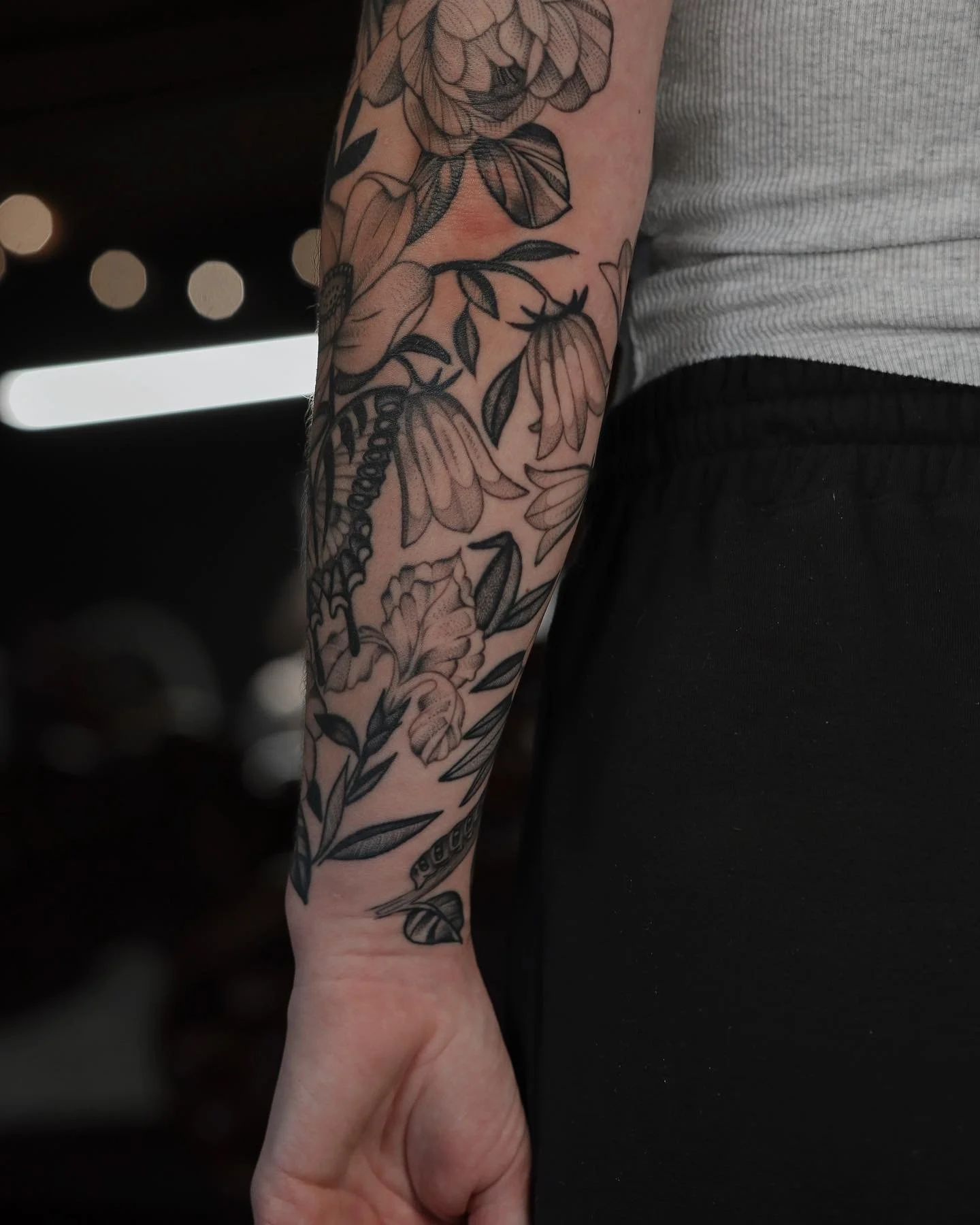 A person's arm with a black and gray floral tattoo covering the forearm. The tattoo includes flowers, leaves, and a caterpillar.