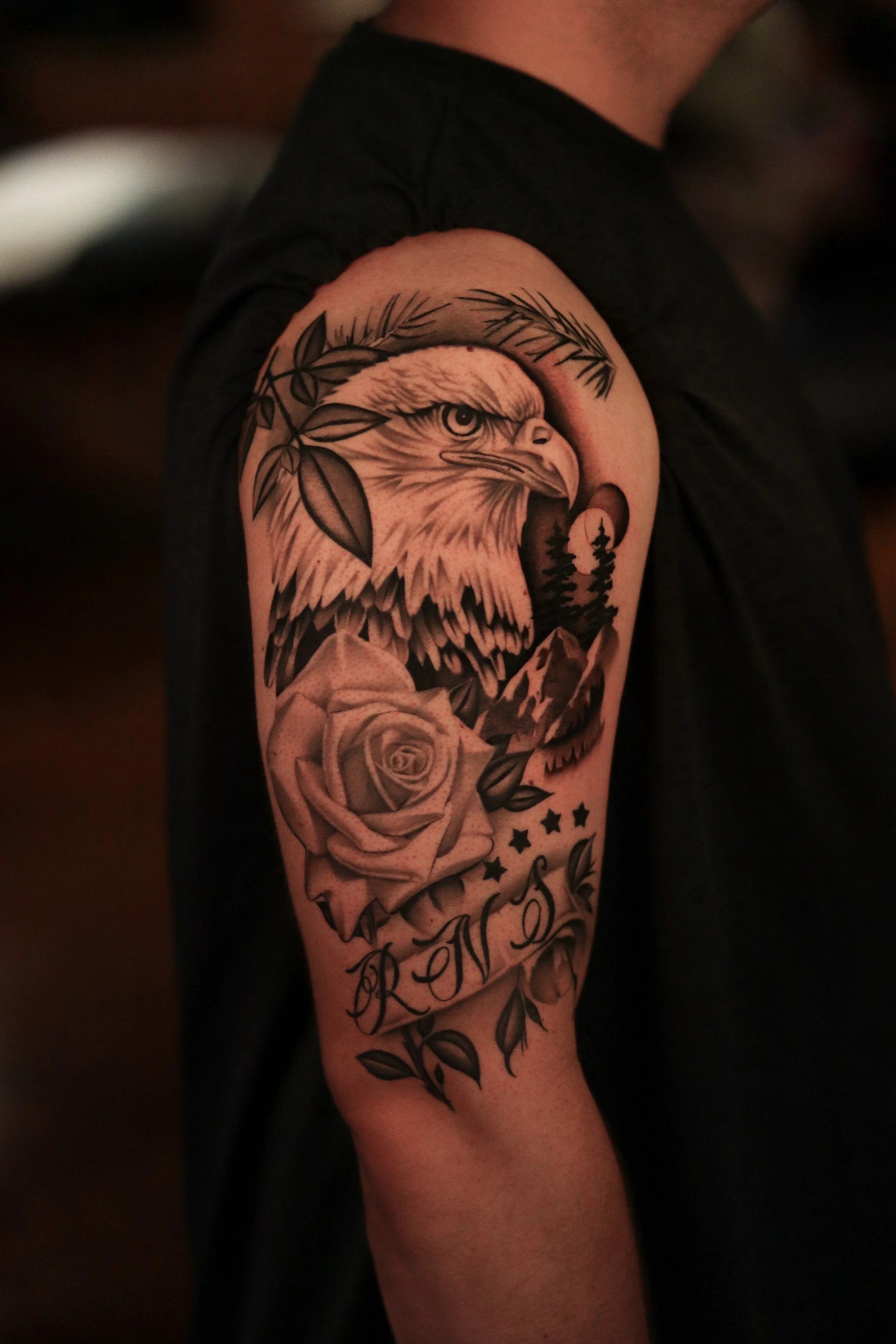 Tattoo of an eagle's head surrounded by leaves, with a rose and a banner with script beneath.