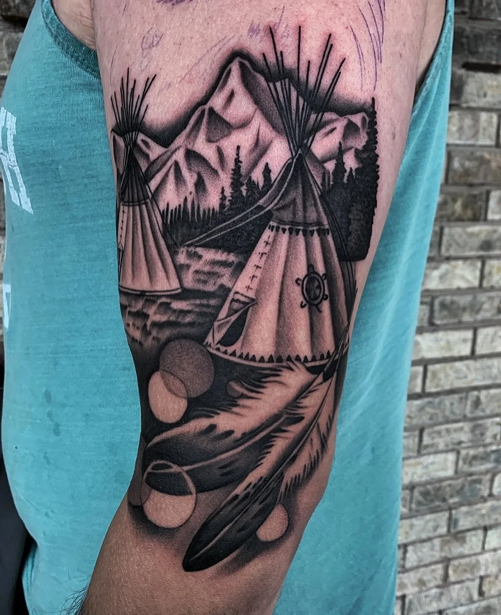 A black and gray tattoo of a mountain landscape with pine trees, mountains, and a river, featuring a teepee with a small knot emblem on it, surrounded by feathers and beads.
