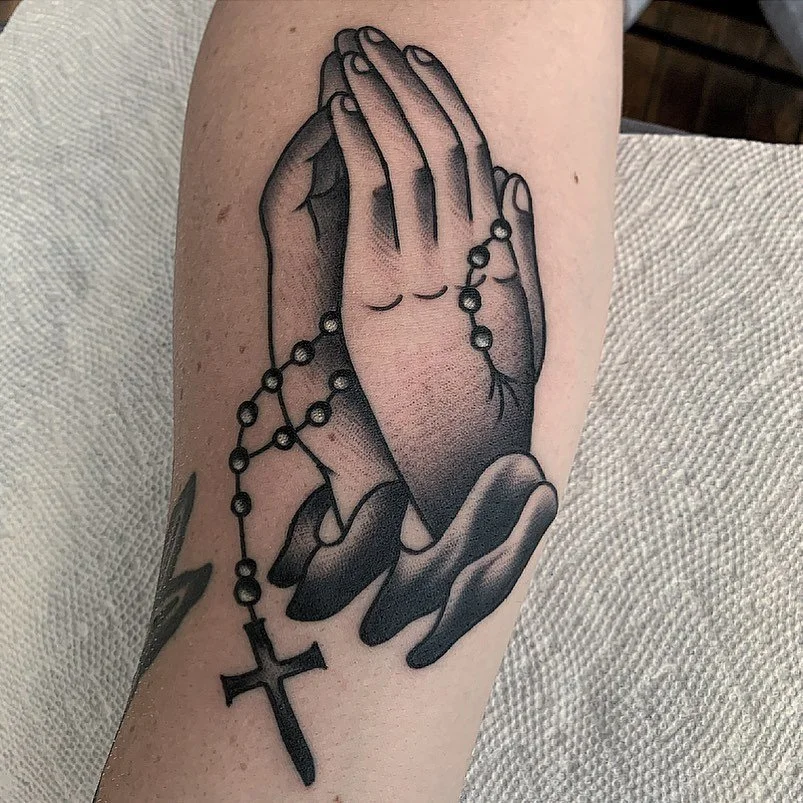 Tattoo of two praying hands holding a rosary with a cross on a person's forearm.
