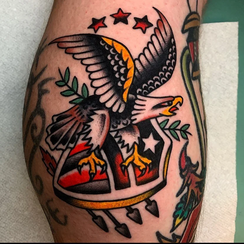 Traditional-style tattoo of a bald eagle with outstretched wings, holding a branch in its talons, surrounded by stars and a banner with hearts hanging from it.