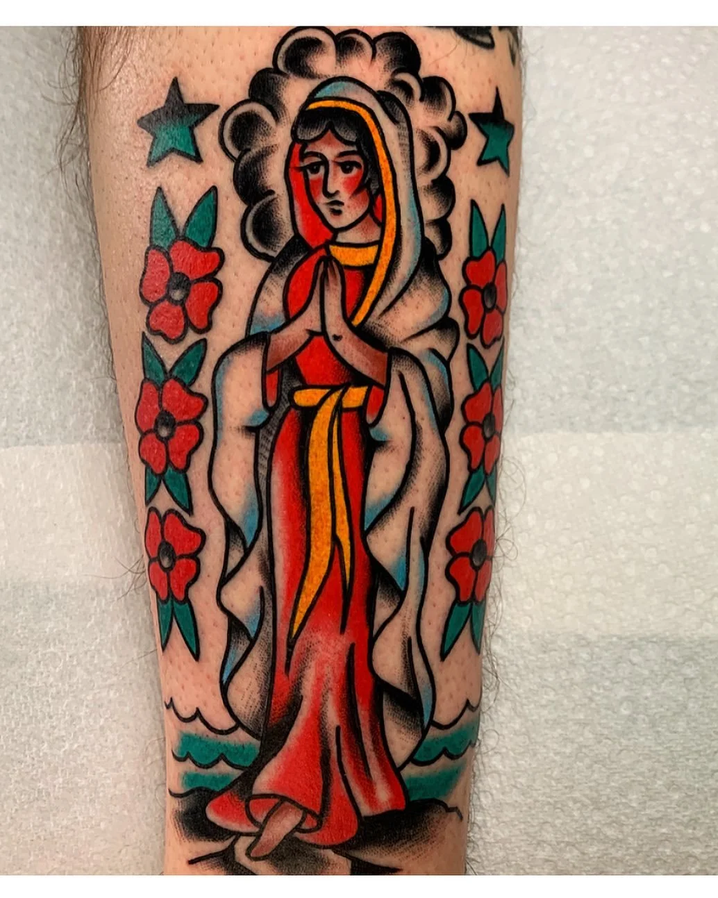 Traditional style tattoo of the Virgin Mary with hands clasped, surrounded by red flowers and teal stars.