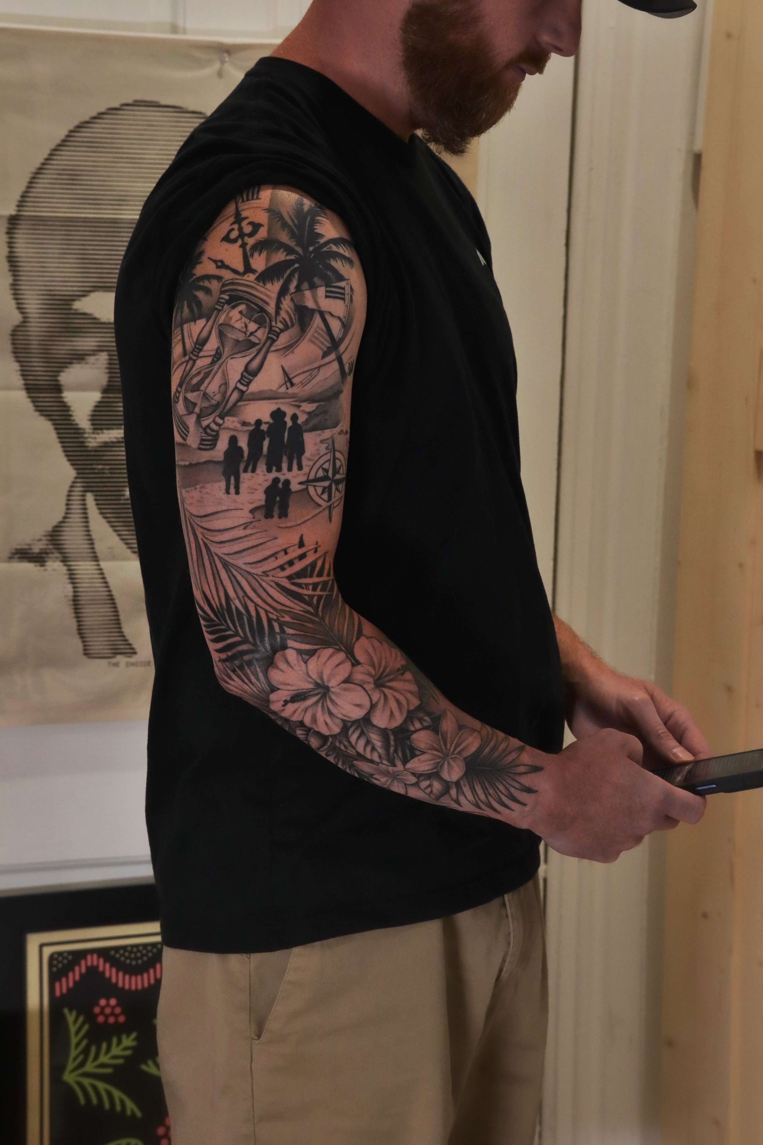 A man with a beard wearing a black sleeveless shirt and beige pants, showing a detailed tattoo sleeve on his left arm with palm trees, a sunset, and a group of people, while holding a phone.
