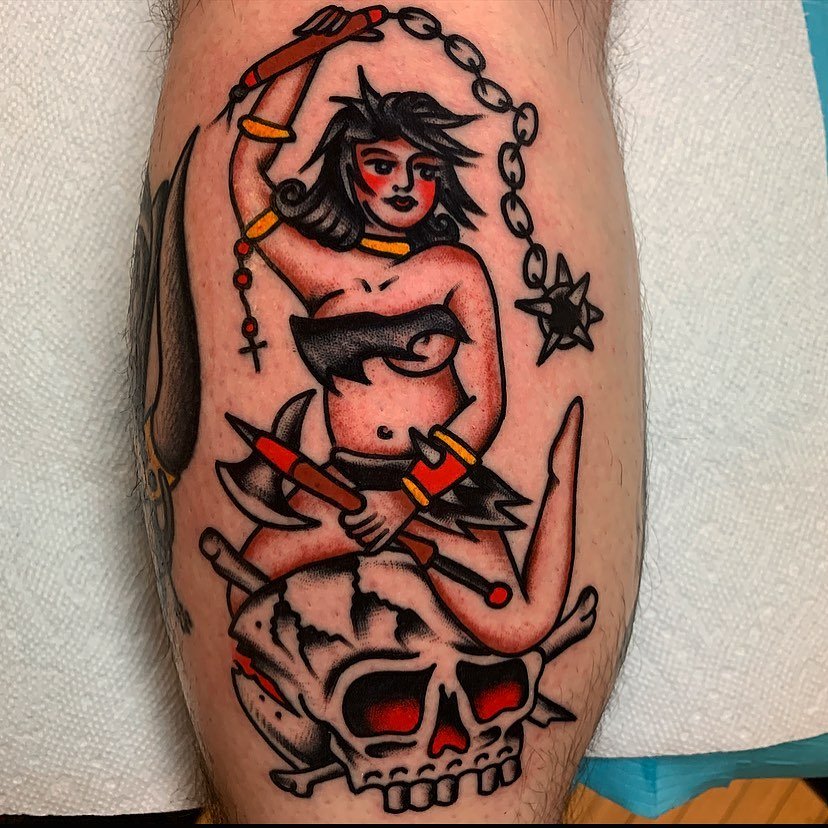Tattoo of a cartoon-style woman with dark hair, holding a chain, sitting on a skull, with a dagger in her hand, wearing minimal clothing, surrounded by accessories like bracelets and a necklace.