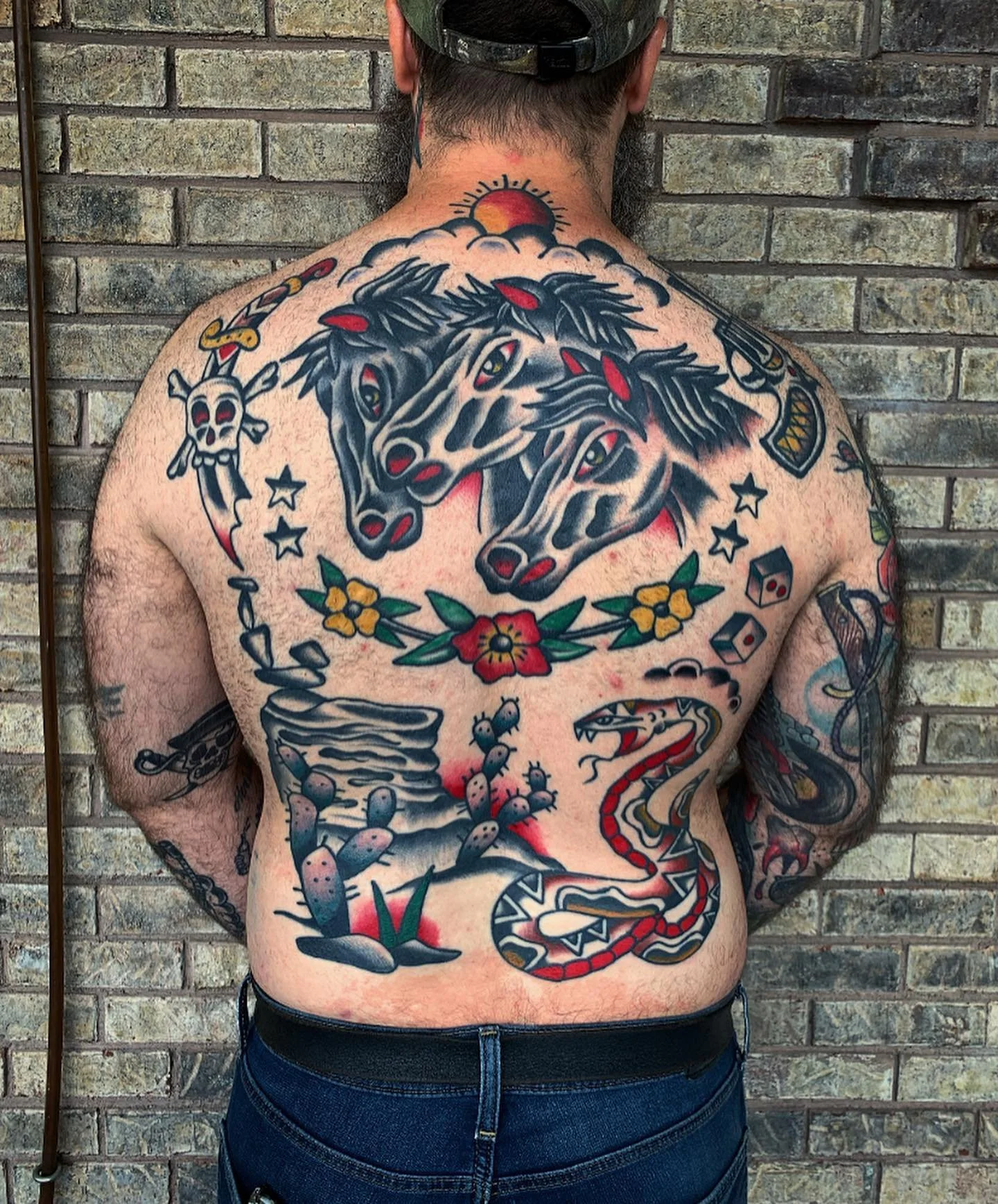 A man with tattoos on his back and arms, featuring a large tattoo of two horses' heads and a snake coiled around his lower body, standing against a brick wall.