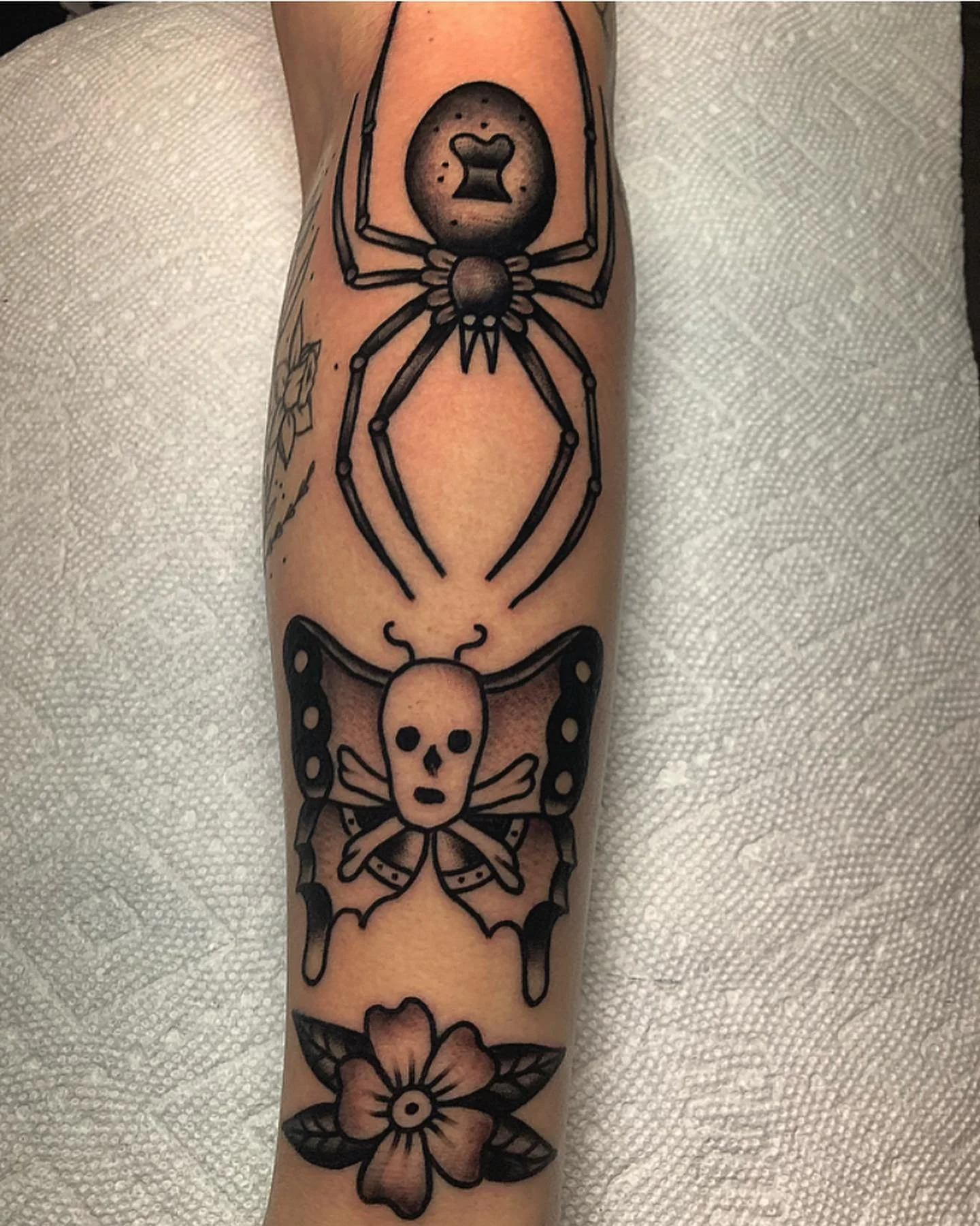 Tattoo of a spider with an oval body containing a small heart-shaped design, on a person's arm. Below the spider, there is a skull with a bow and wings, and a large flower at the bottom.