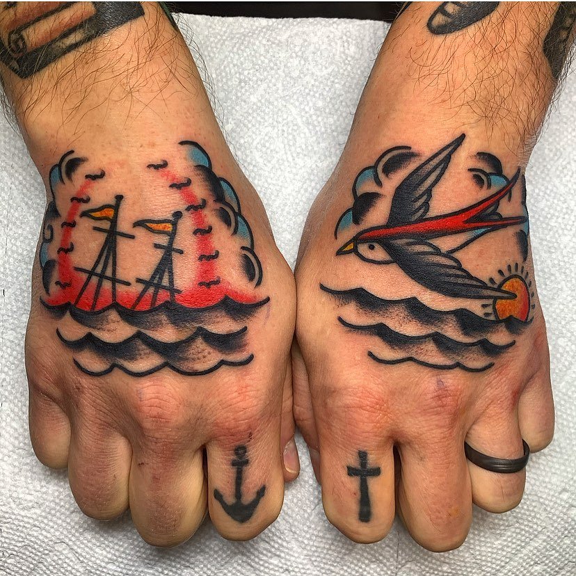 Tattooed hands with nautical-themed tattoos of ships, a swallow, an anchor, a cross, and a sunset over water.