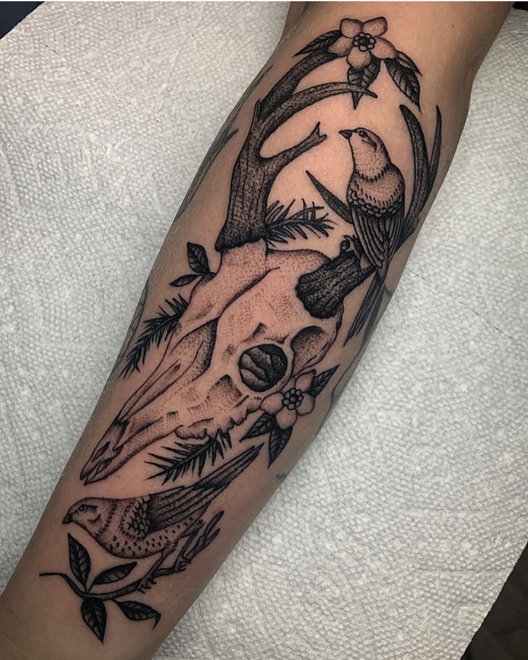 Black and grey tattoo of a deer skull, birds, flowers, and leaves on a person's forearm.