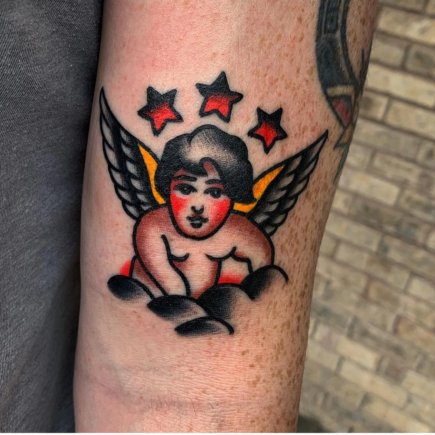 Tattoo of a woman with dark hair and red cheeks, with black wings, surrounded by three red and black stars, on a person's arm.