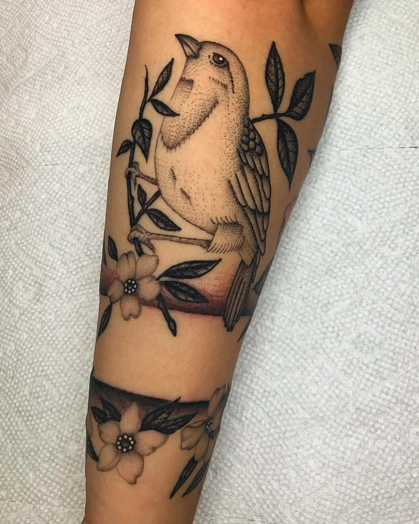 Tattoo of a bird perched on a branch with leaves and flowers on someone's arm.