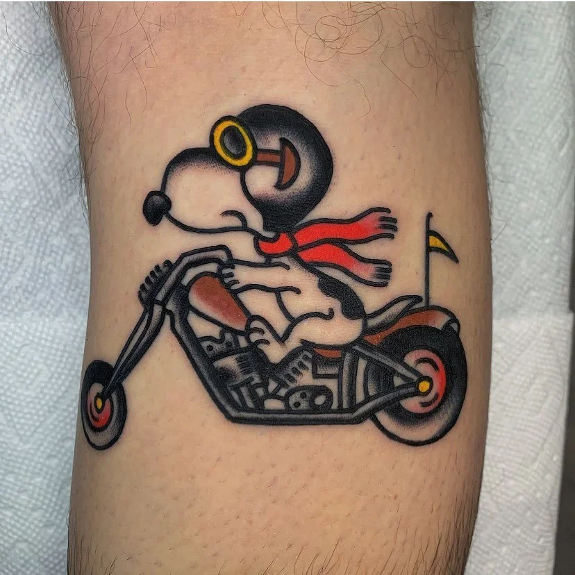 A cartoon tattoo of Snoopy as the Flying Ace riding a motorcycle, wearing aviator goggles, a red scarf, and a helmet, with a small flag attached to the motorcycle.