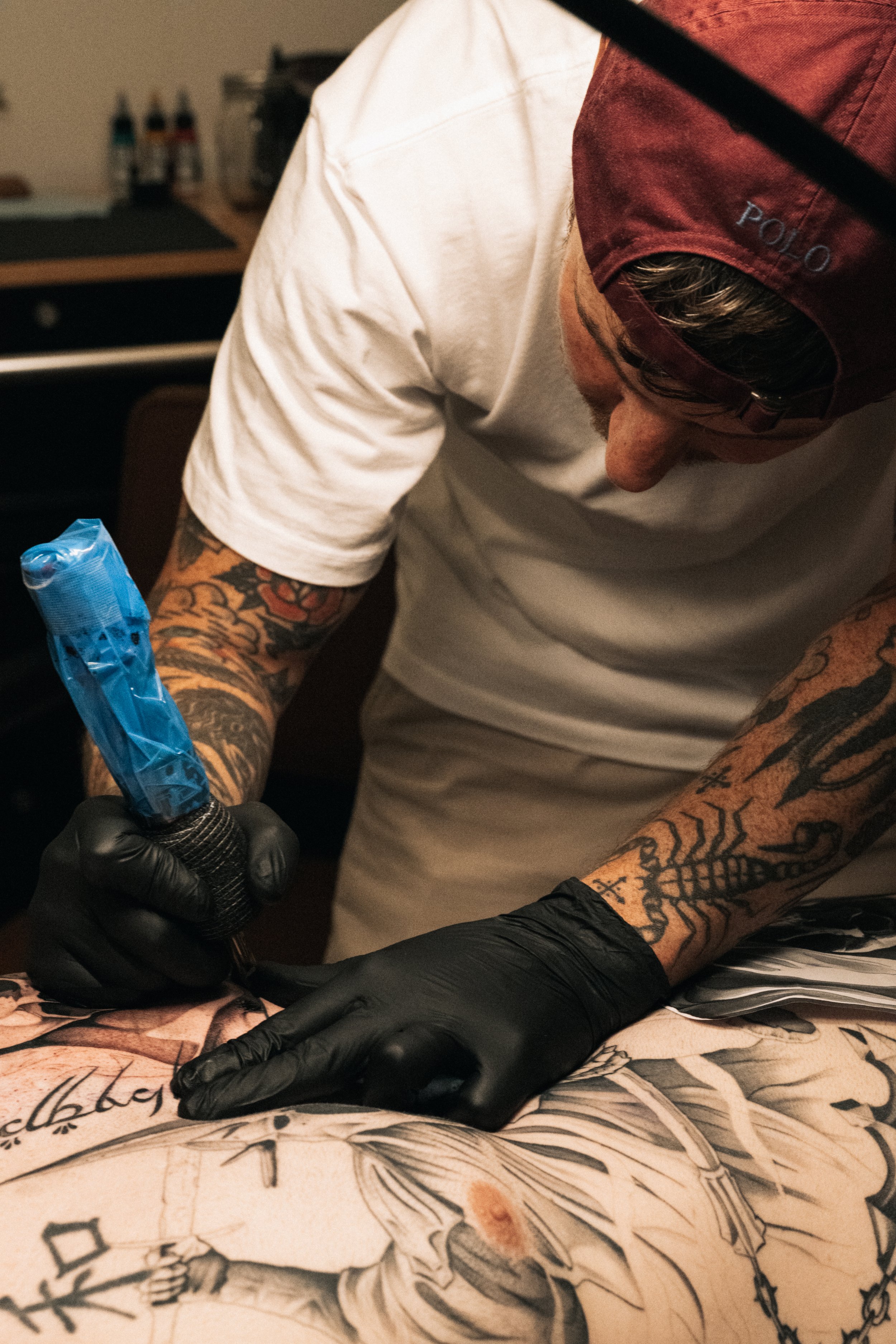 A person with tattoos on their arms, wearing a red Polo baseball cap backwards, working on a tattoo with black gloves, using a tattoo machine. The tattoo appears to be a detailed line drawing on skin.