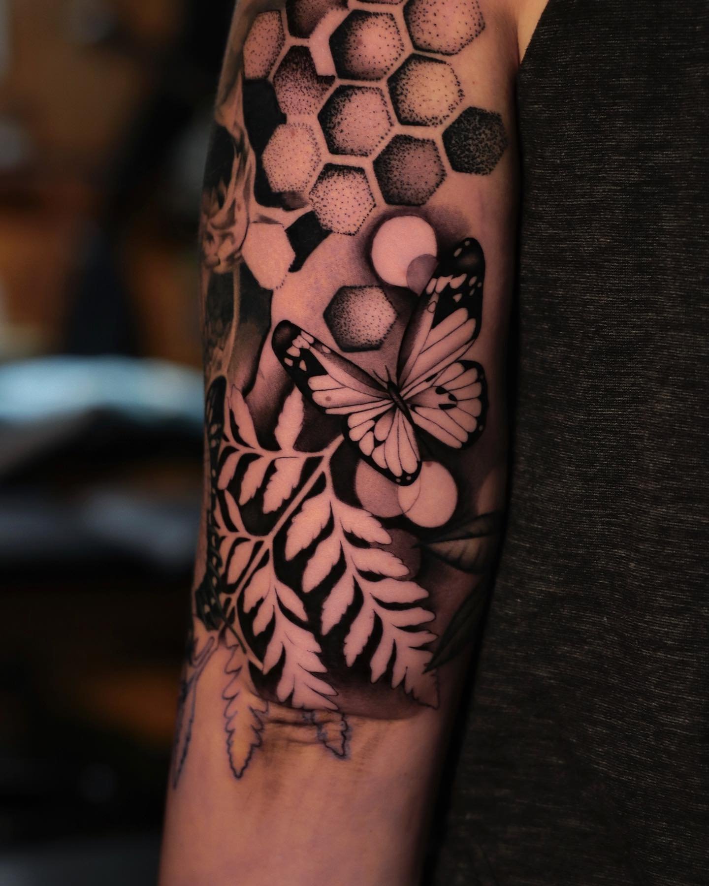 Black and gray tattoo of a butterfly, honeycomb pattern, and leaves on person's upper arm.