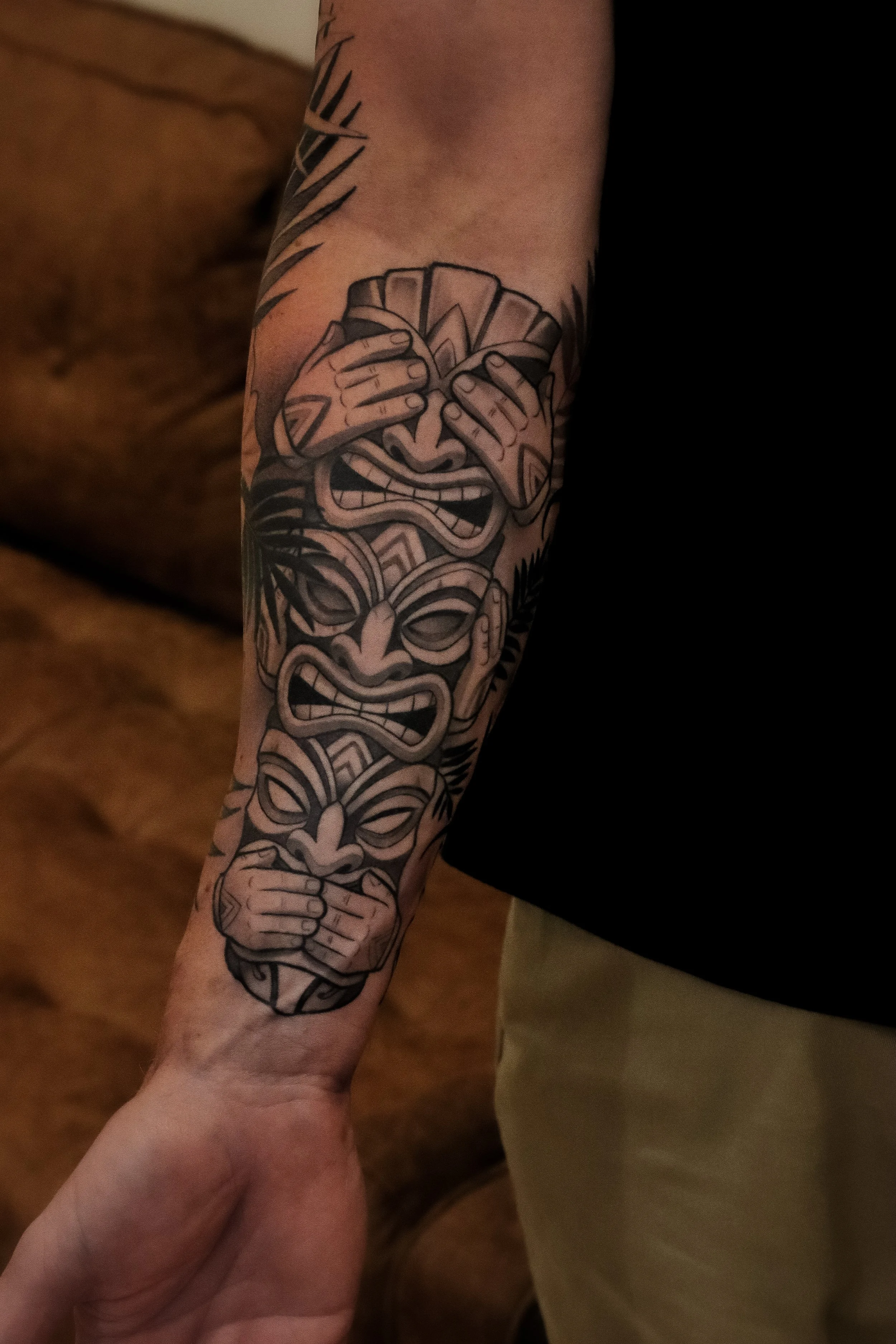 A tattoo on a person's arm depicting a tiki mask and a person holding a fan.