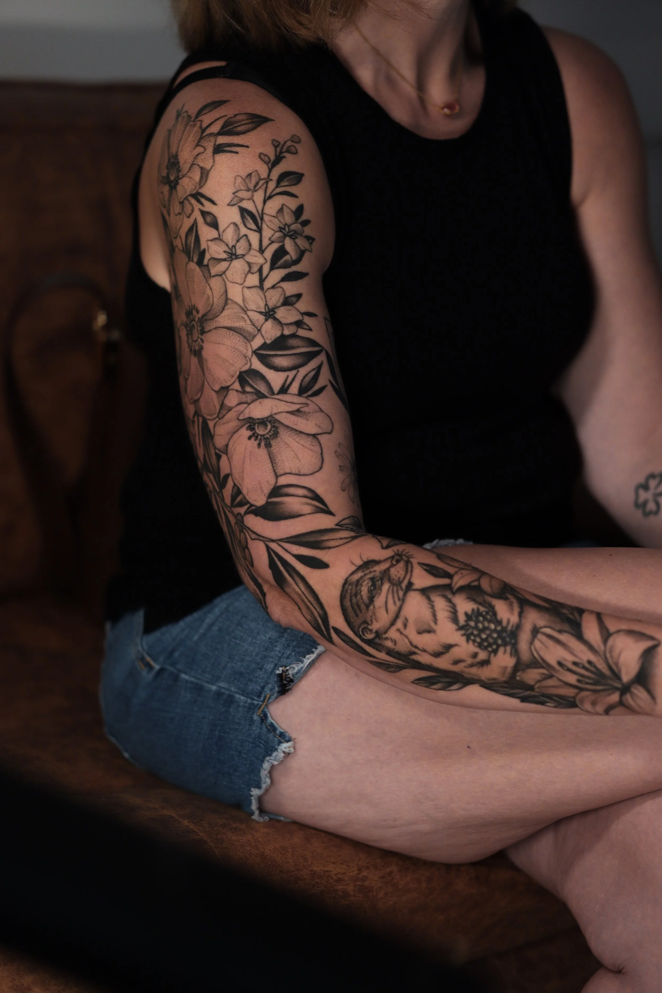 A woman wearing a black tank top and denim shorts with tattoos on her arm and thigh, sitting on a wooden surface.