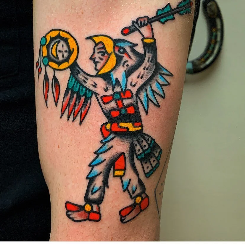 Tattoo of a stylized Native American warrior with feathers, a bow and arrow, holding a shield, and wearing traditional clothing with red, black, and blue accents.