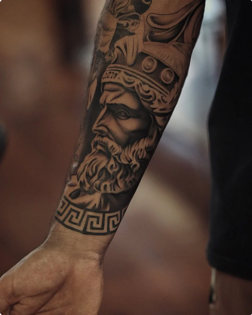 Tattoo of a bearded man wearing a helmet with decorative details, set on the inner forearm.