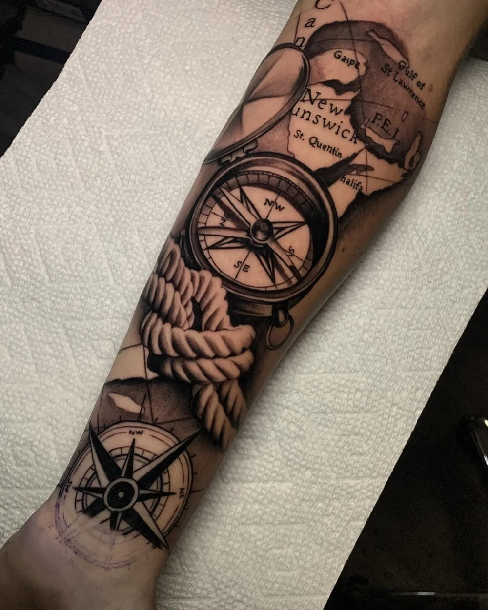 Tattoo of a map of New Brunswick, Canada, with a compass and a rope coiled around a compass on a person's forearm.
