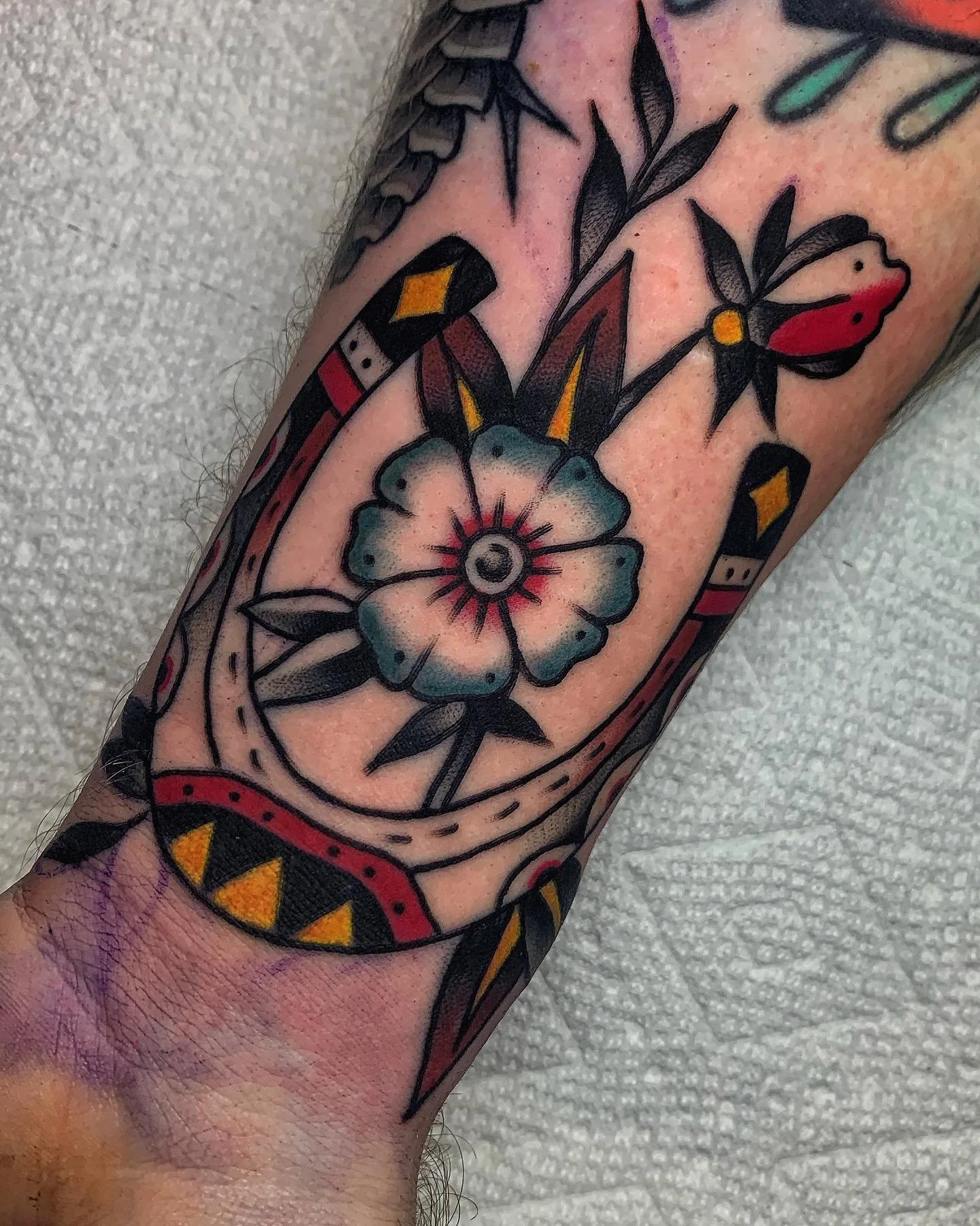 Close-up of a tattoo on a person's arm featuring a colorful flower with a blue hue, black outline, and a red center, along with decorative geometric and floral elements surrounding it.