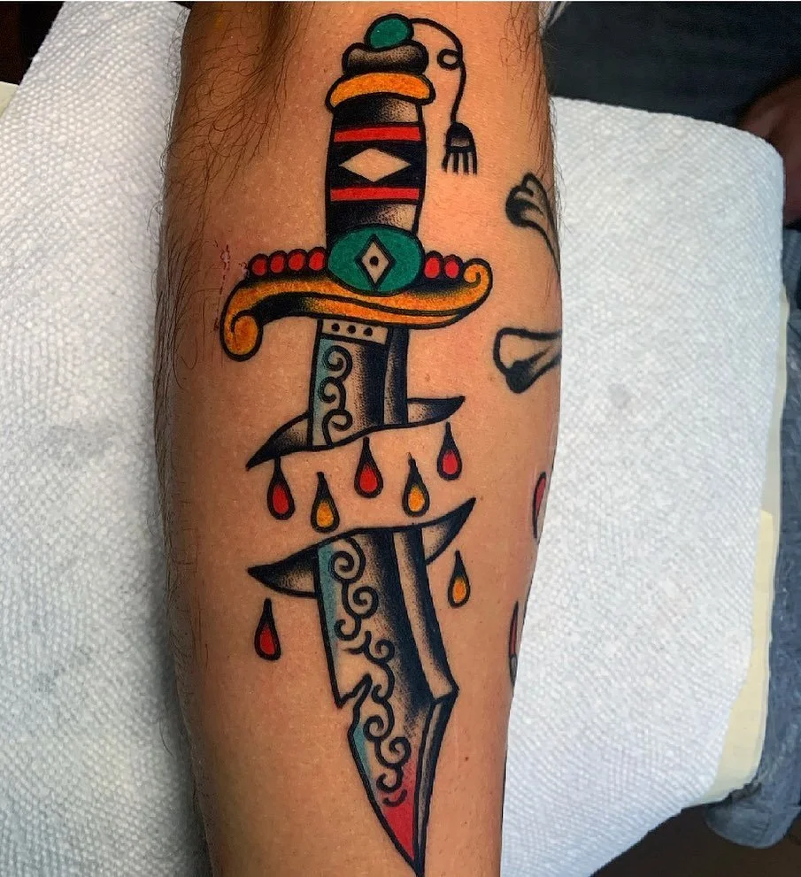 Tattoo of a colorful sword with red, black, yellow, green, and blue details, featuring teardrop-shaped red and yellow drops hanging from the crossguard.