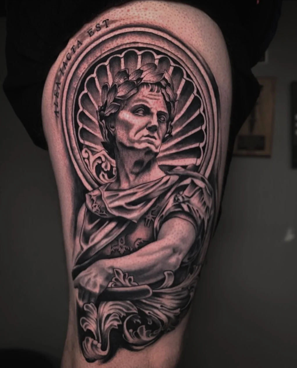 Black and gray tattoo of a classical statue with a laurel wreath, surrounded by a circular frame with rays, and the words 'NEBACH EST' above.