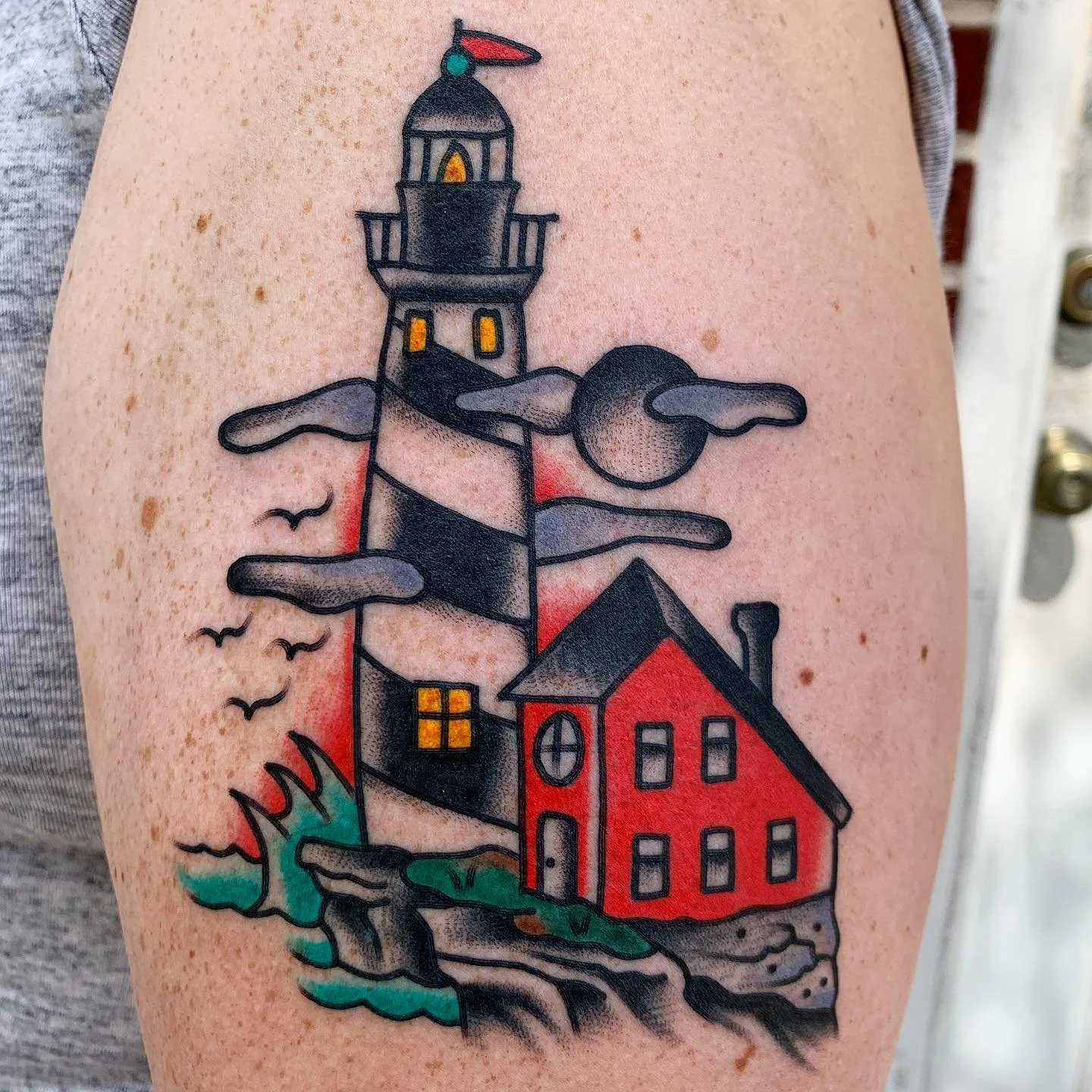 Colorful tattoo of a lighthouse with a red house at its base, clouds and seagulls, and water at the bottom.