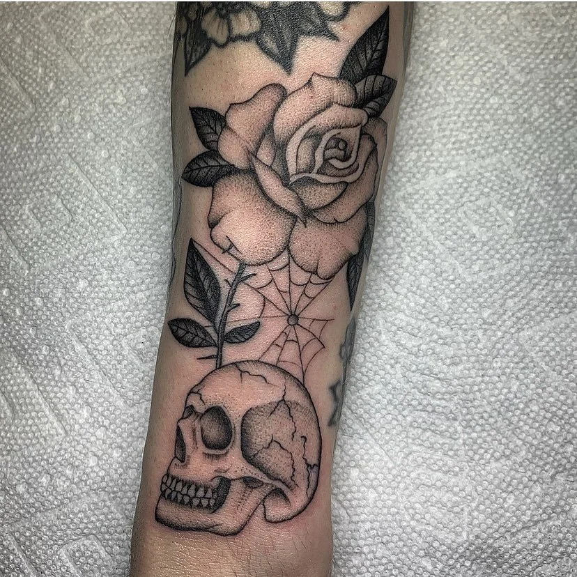 Tattoo of a rose flower, leaf, spider web, and skull on a person's arm.
