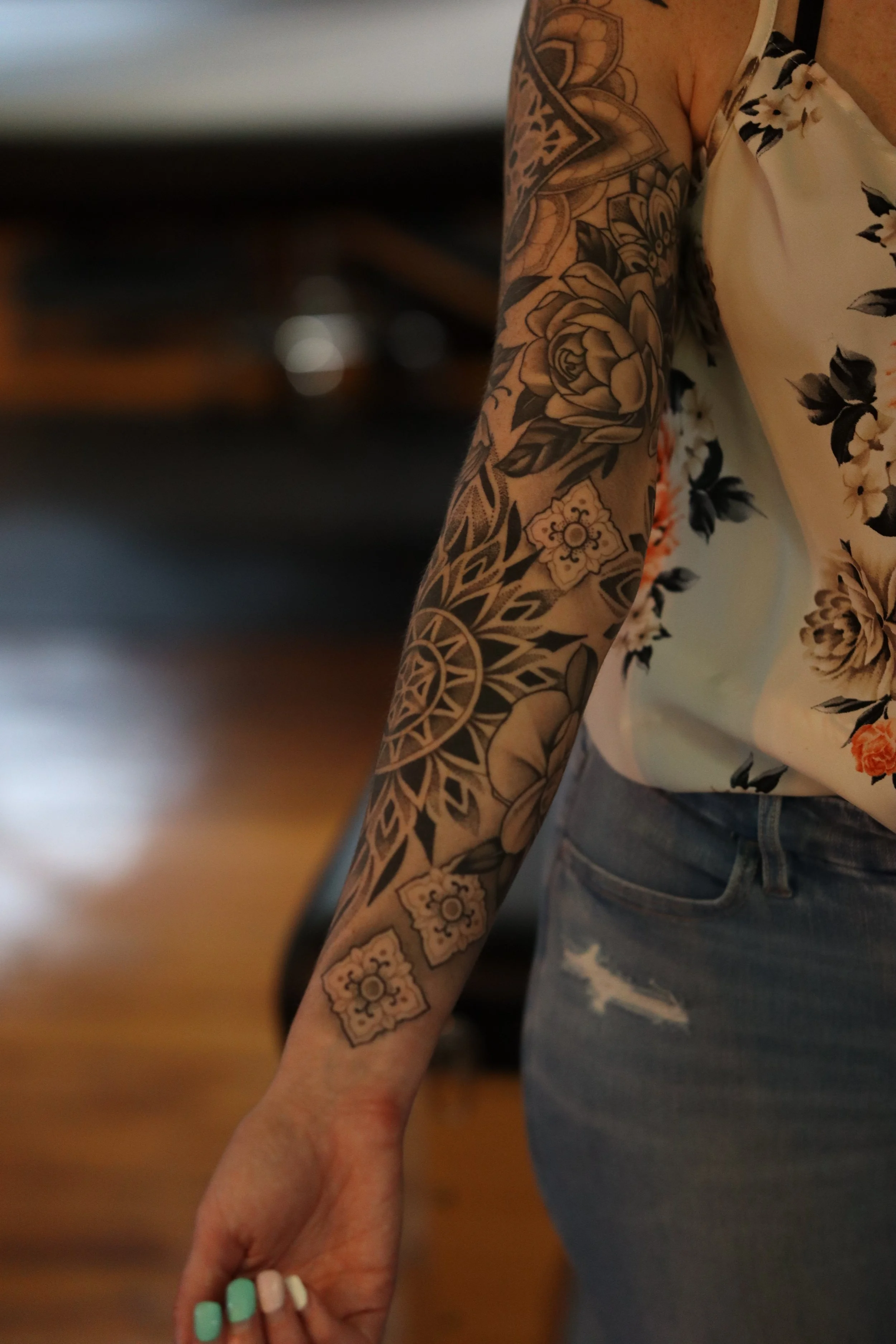 A person with a floral tattoo on their arm and wearing floral-patterned top, holding a hand with painted nails.