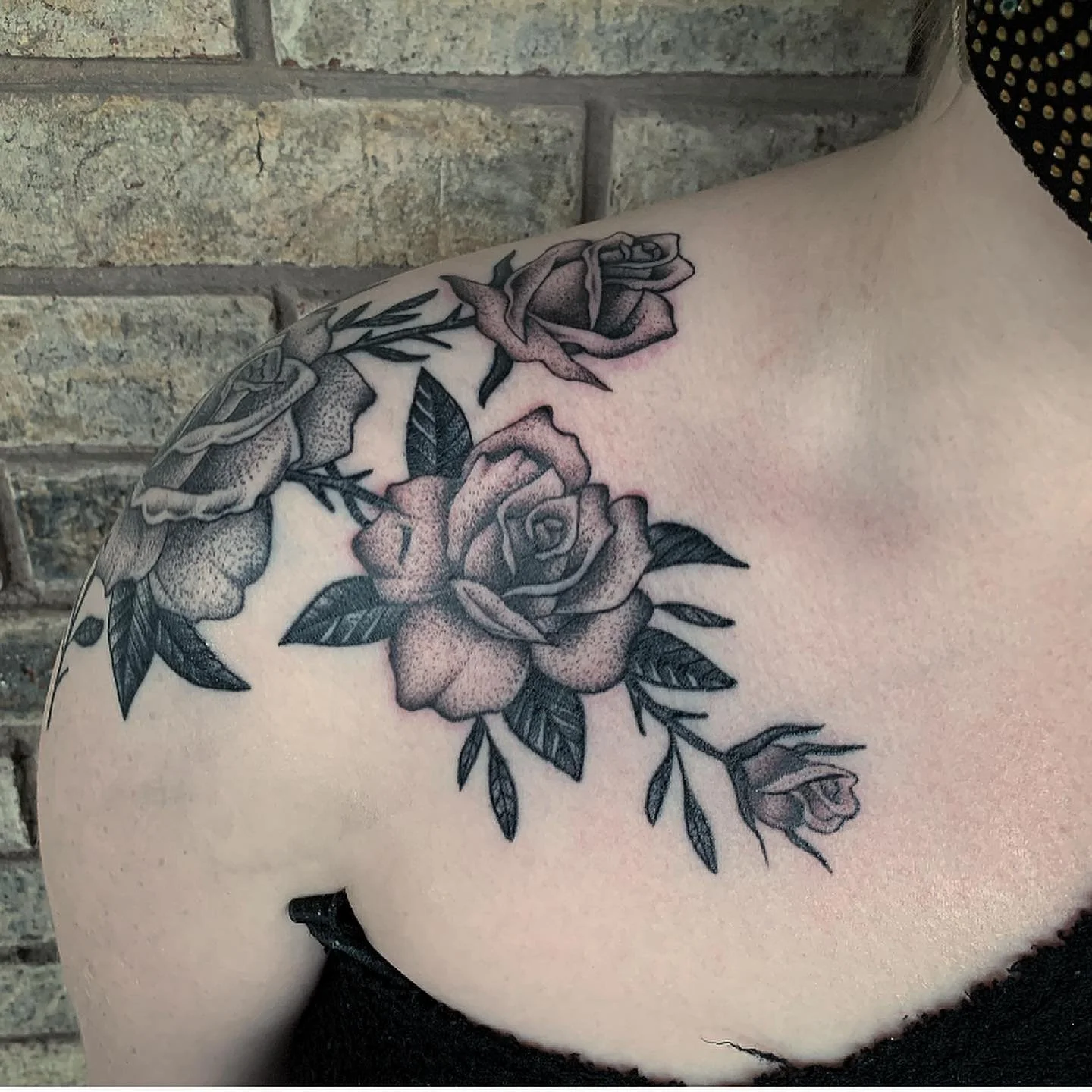 A black and gray floral tattoo featuring roses and leaves on a person's shoulder and upper arm, with a brick wall in the background.