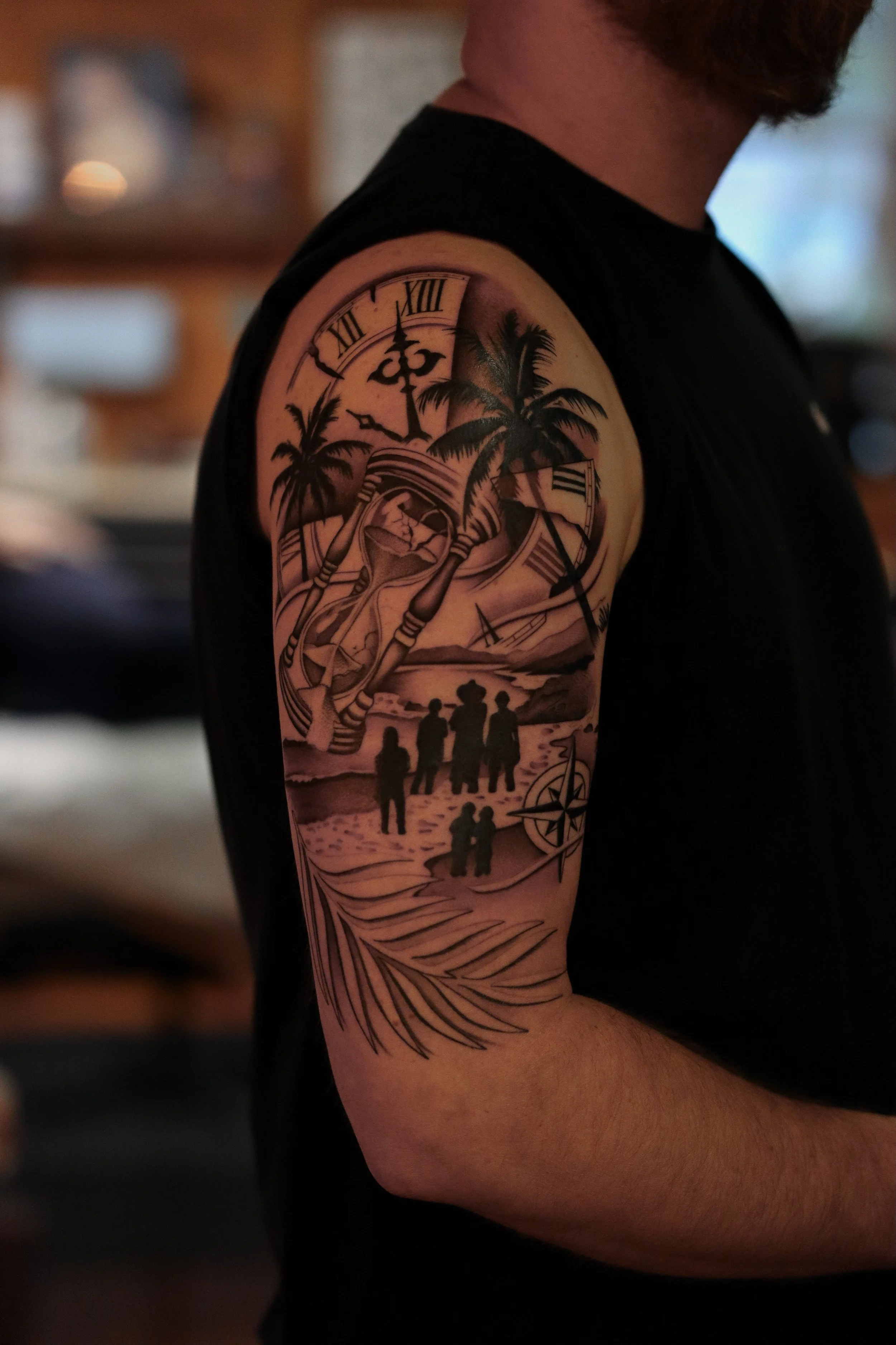 Tattoo of a beach scene with palm trees, a sunset, a compass, an hourglass, and a silhouette of a family walking, on a man's upper arm.