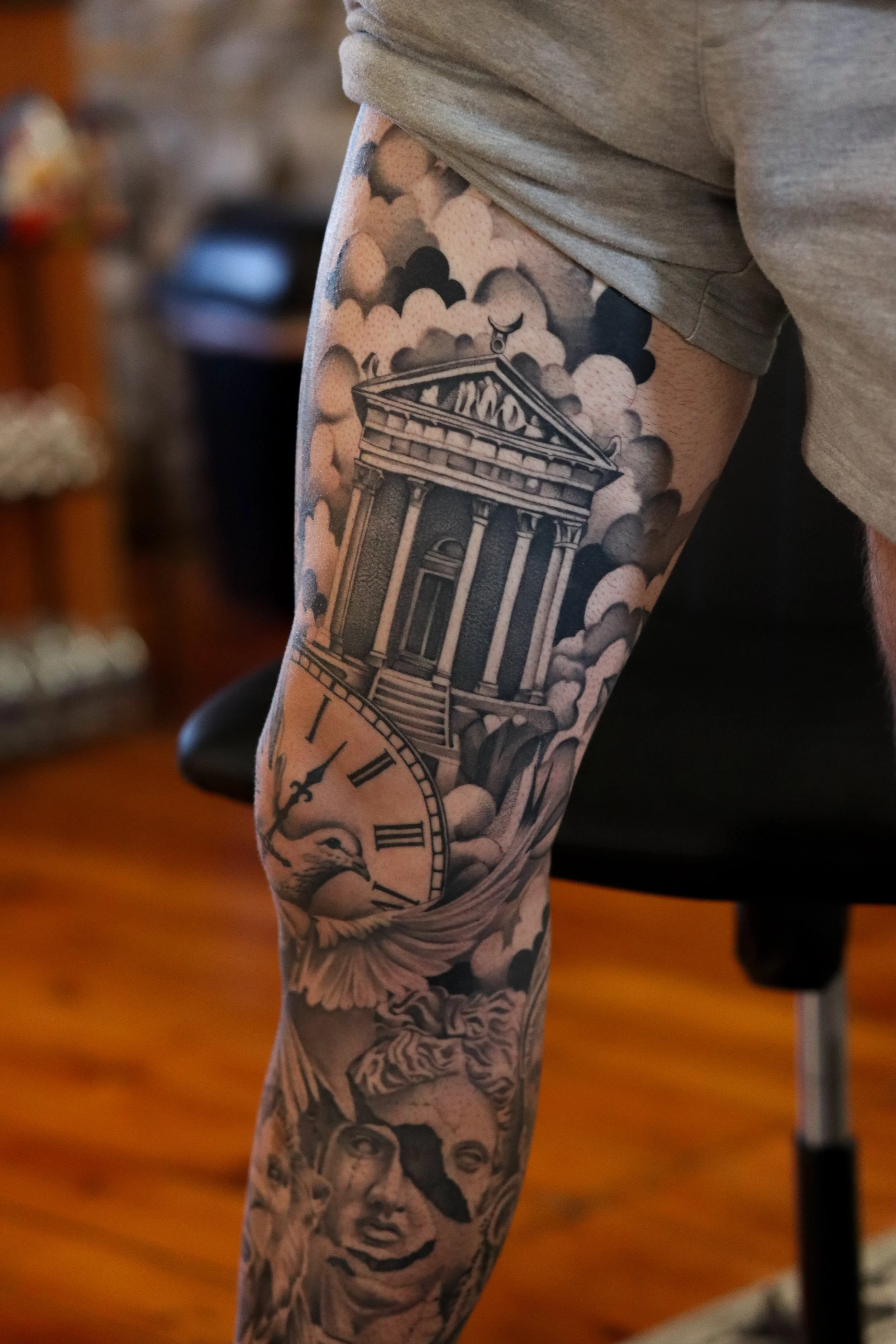 Close-up of a person's leg covered in detailed black and gray tattoos, including a classical building, a clock, a woman's face, and clouds.