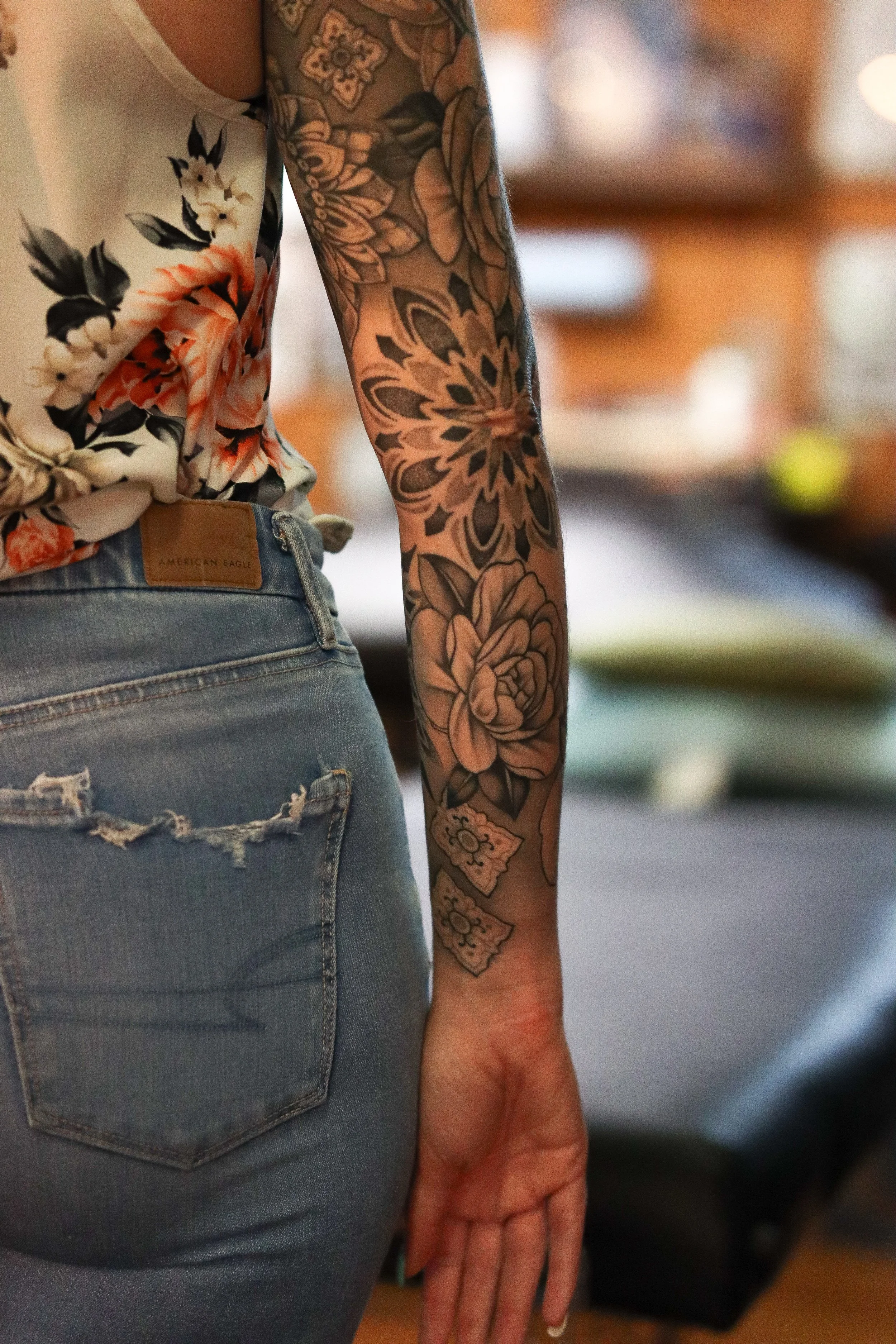 A person wearing a floral blouse and ripped jeans with a leather patch that says 'American Eagle.' The person's arm has a large, detailed black and gray floral tattoo.