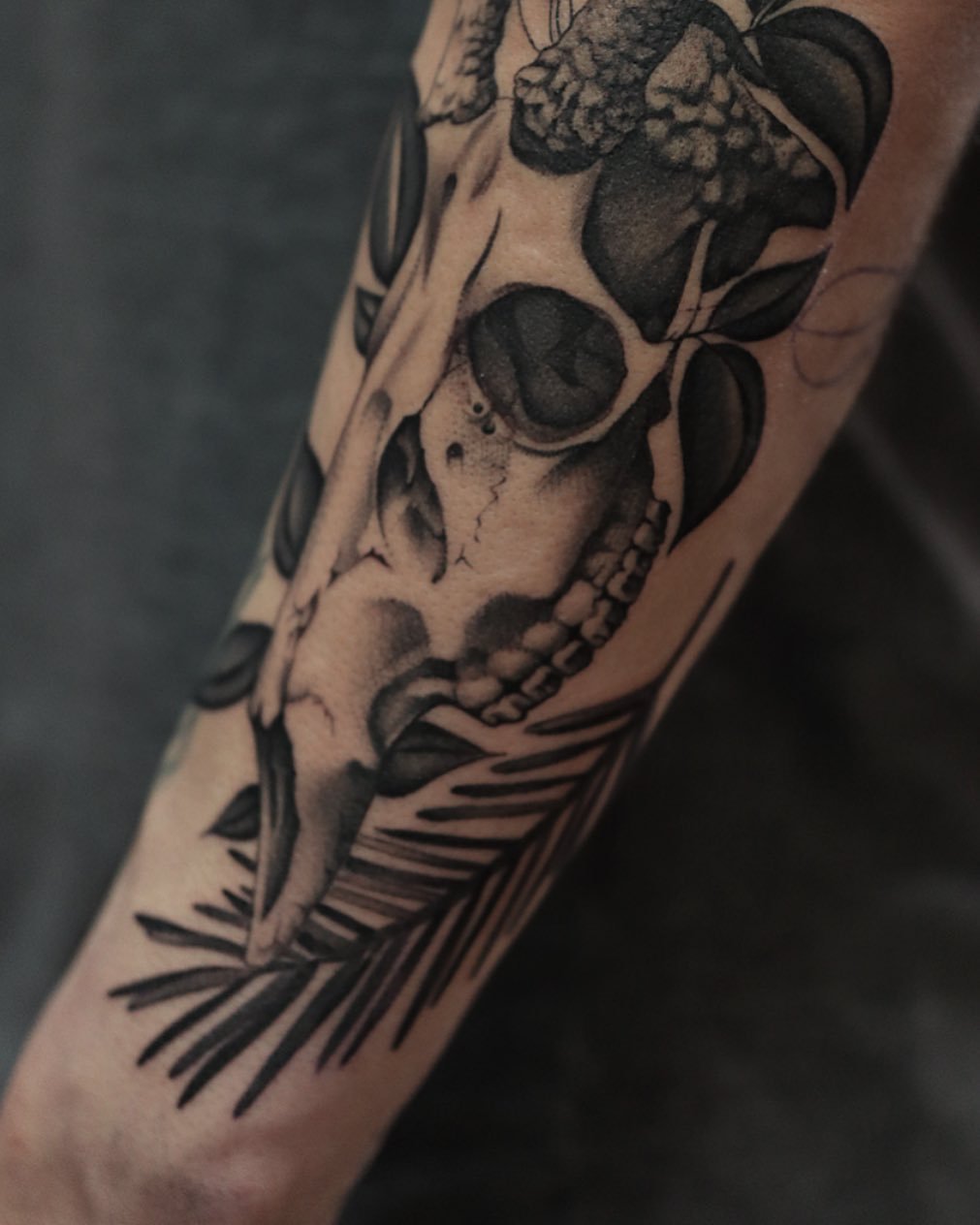 A black and gray tattoo of a human skull with floral elements and leaves on a person's forearm.