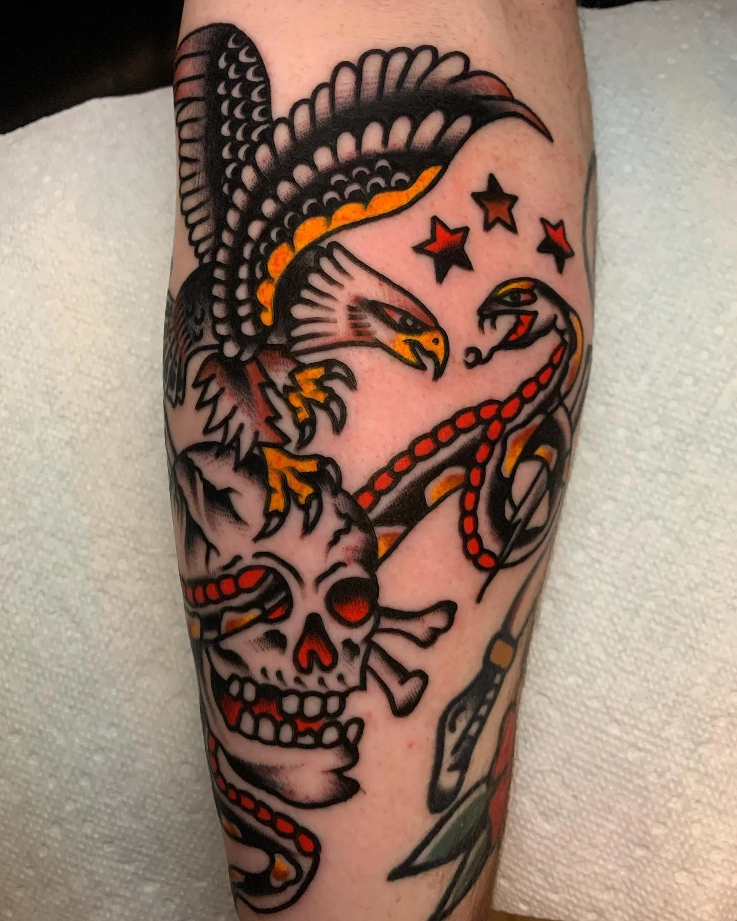 Tattoo featuring a skull, an eagle, a snake, three stars, and various decorative elements in black, red, yellow, and orange colors.
