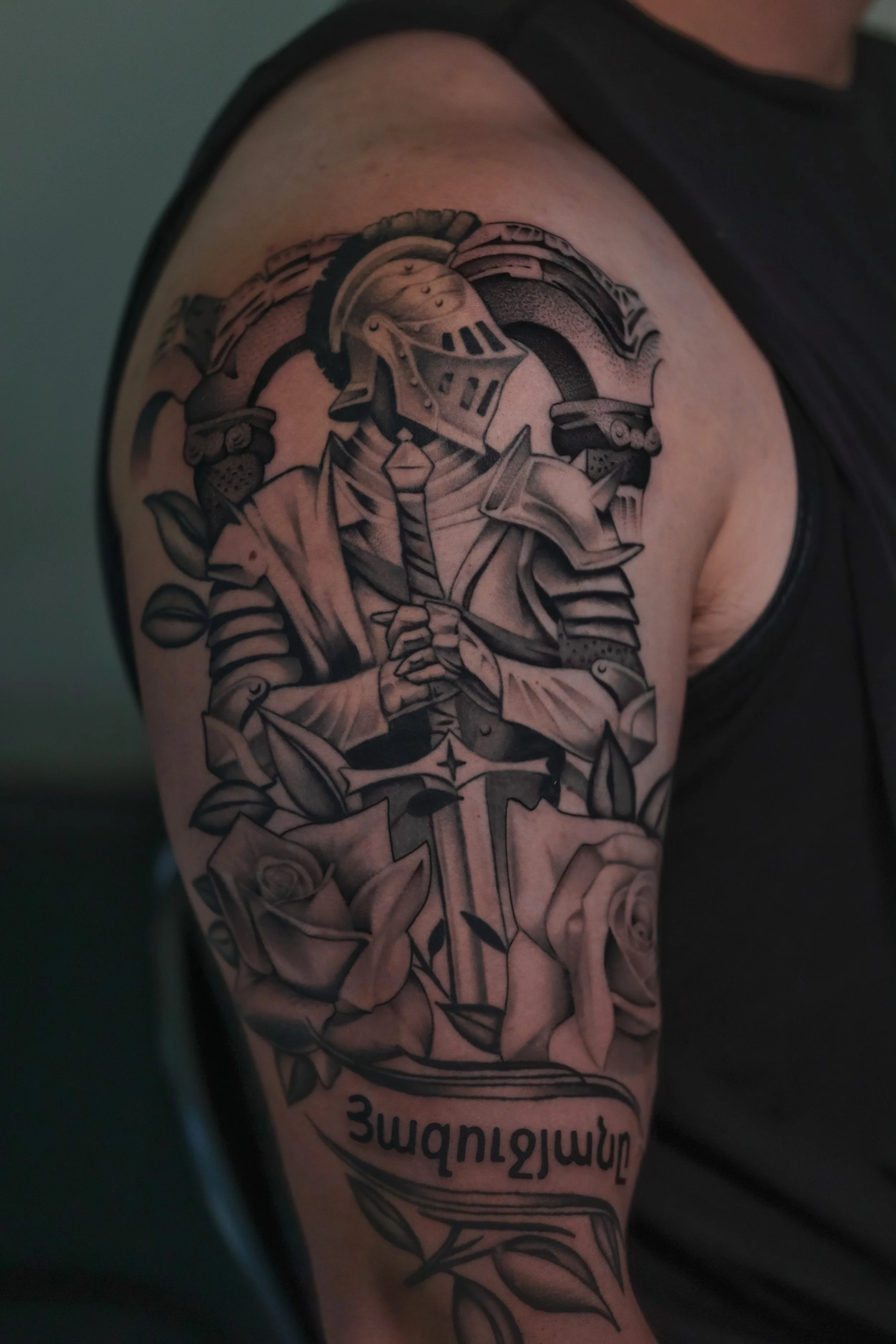 Tattoo on upper arm featuring a knight or warrior with armor, holding a sword, surrounded by roses, with text in a banner underneath.